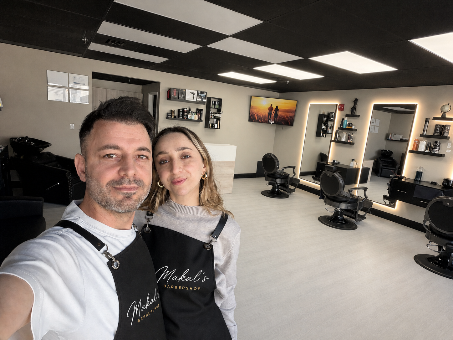 A man and woman taking a selfie inside a modern barbershop with black chairs, mirrors, a television, and shelves with barber supplies.