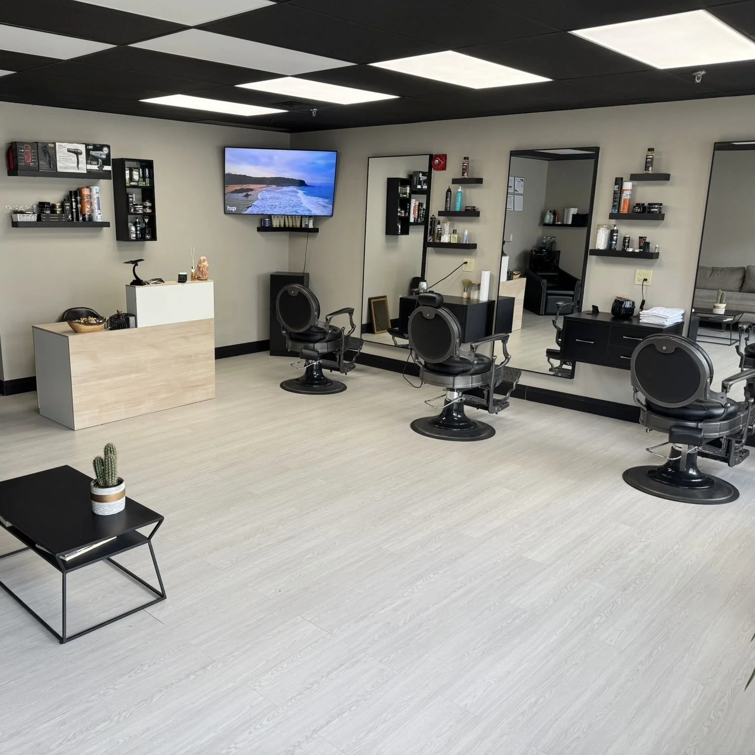 A modern hair salon with three black barber chairs facing a large mirror on the wall. There are shelves with hair products, a small black table with a cactus plant, and a white reception counter. A flat-screen TV displays a landscape, mounted on a beige wall.
