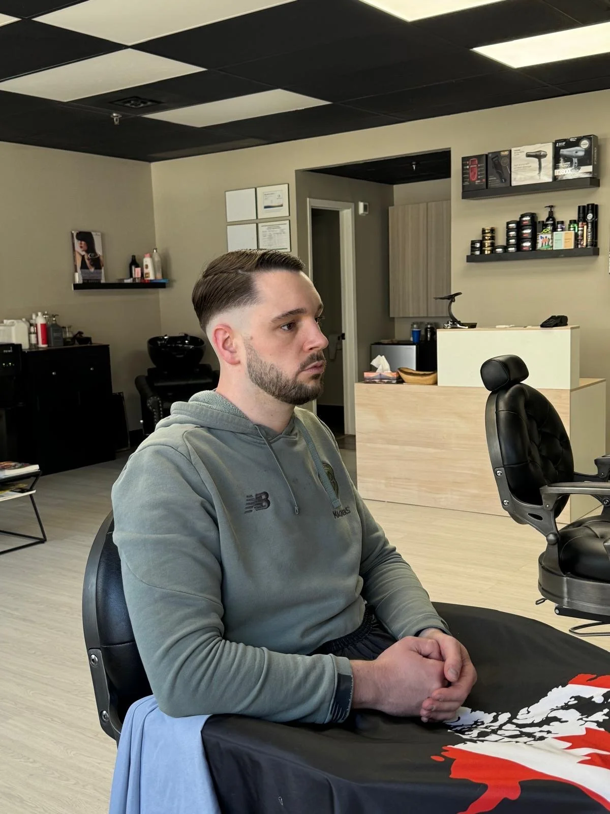 A man with short dark hair and a beard sitting in a salon or barbershop, wearing a gray hoodie, with a hair salon chair nearby and shelves with hair products in the background.