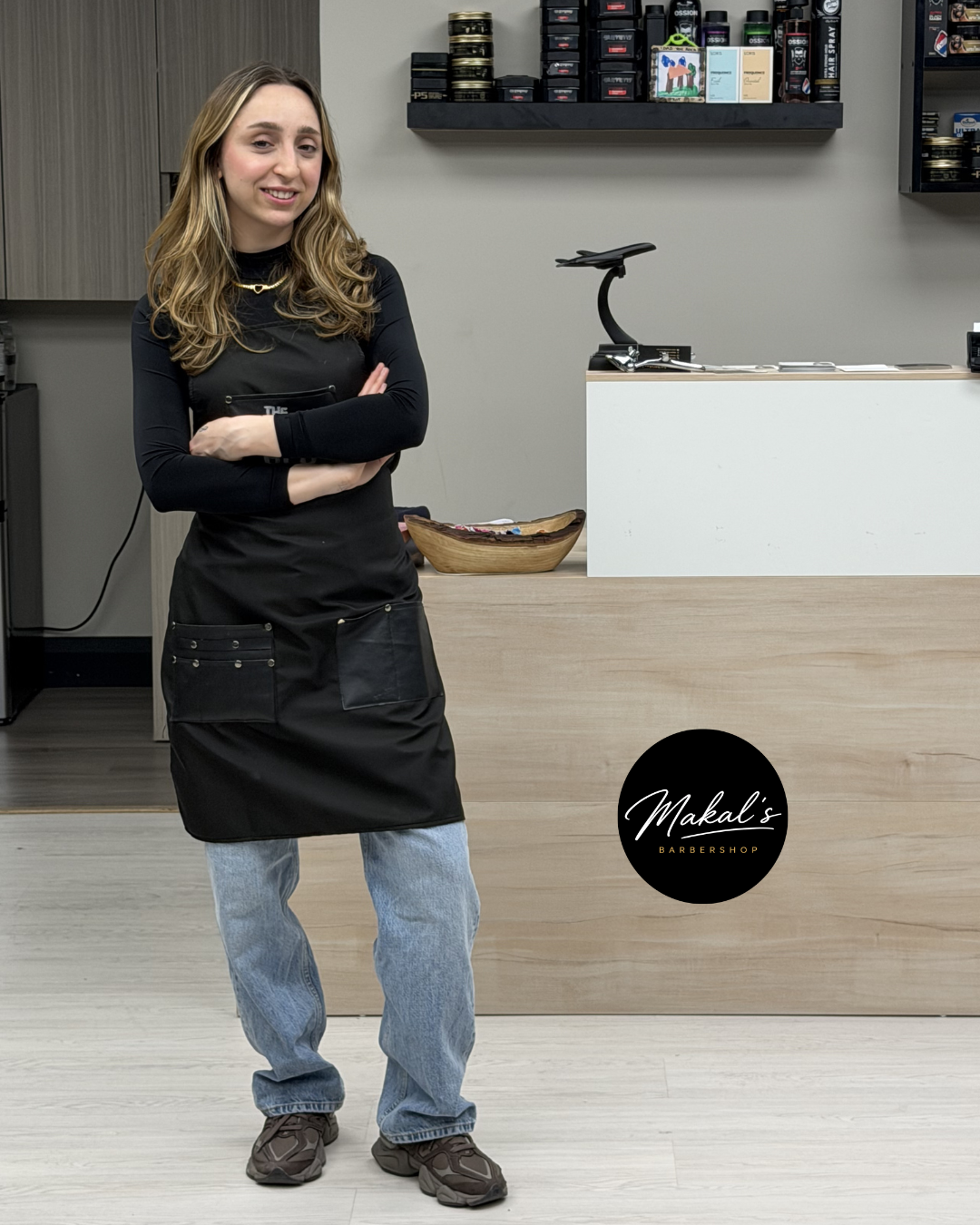 A woman with long, wavy blonde hair standing with crossed arms in a barbershop. She is wearing a black apron, light blue jeans, and sneakers. The barbershop has a light wood counter, shelves with hair products, and a black circular sign that reads "Makal's Barbershop."