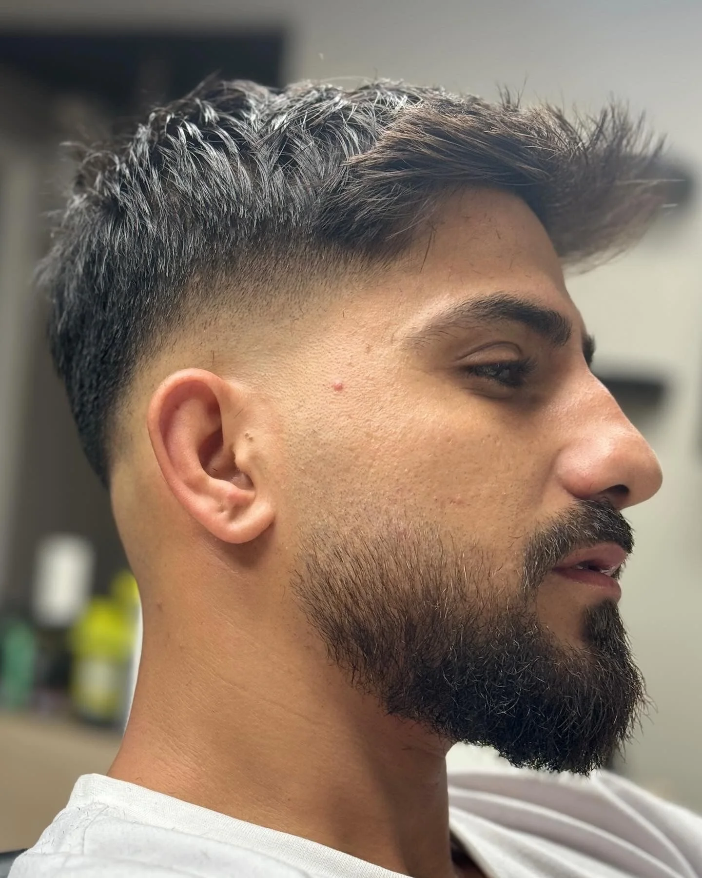 Close-up side profile of a young man with a modern fade haircut and a full beard, wearing a white shirt.