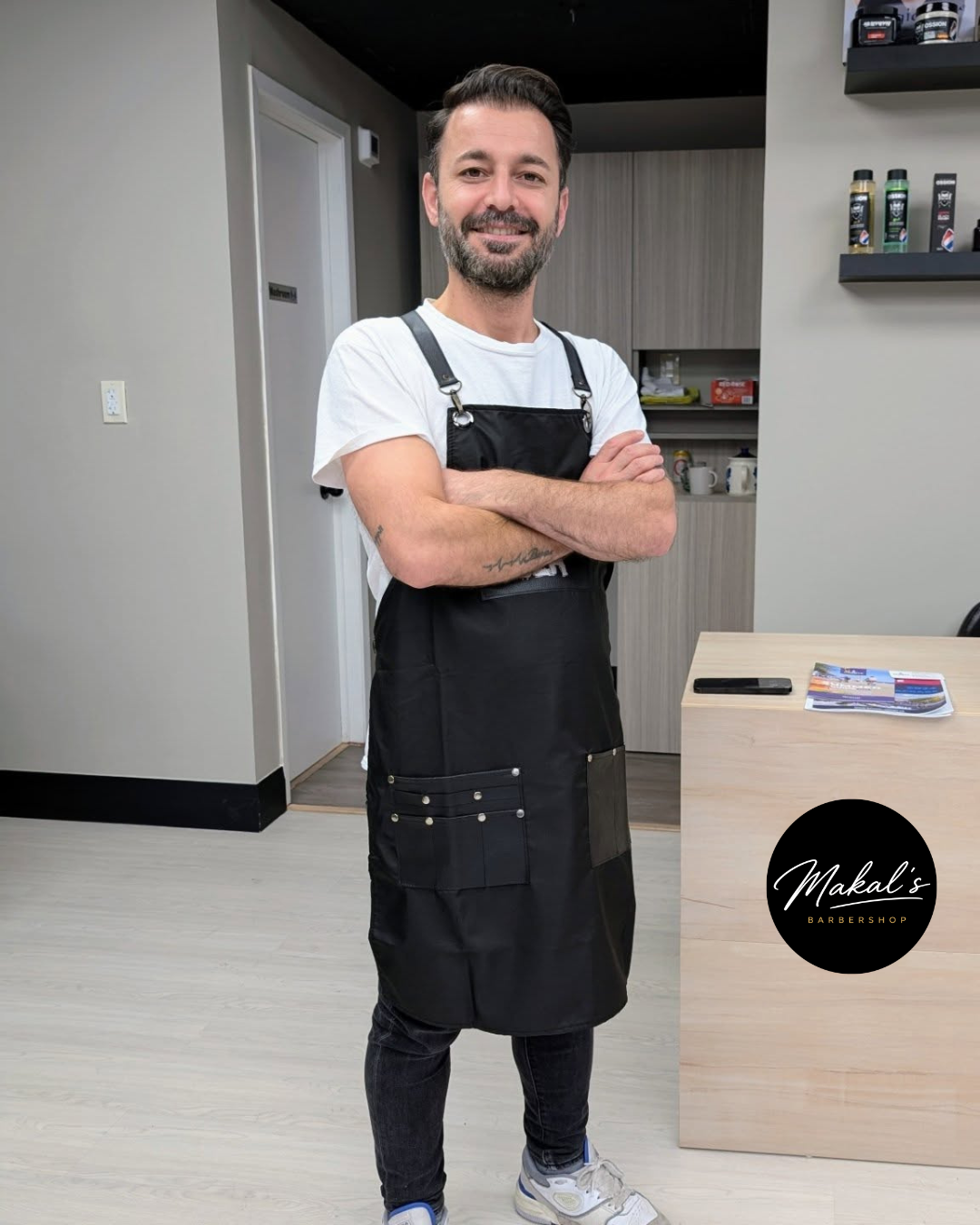 Man with beard smiling, standing with arms crossed, wearing a white t-shirt and black apron, in a modern barbershop. There is a round logo on the table that says "Makal's Barbershop."