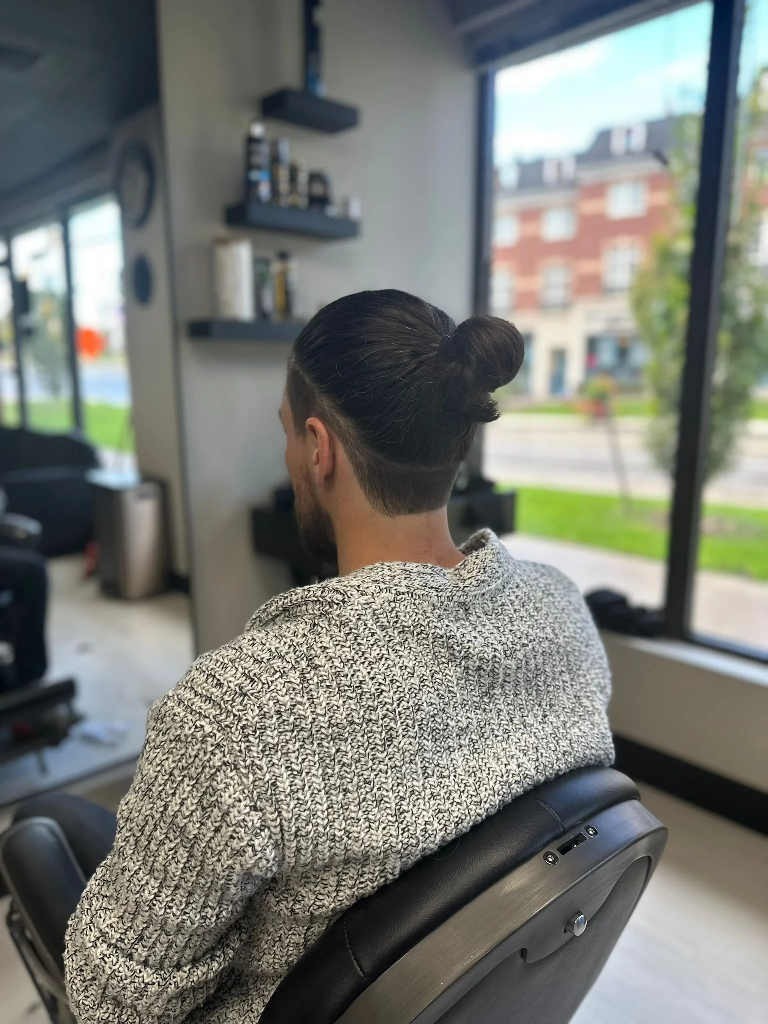 A man with dark hair styled in a bun sitting in a barber chair at a modern barbershop, wearing a patterned sweater, with large windows showing a cityscape of buildings outside.