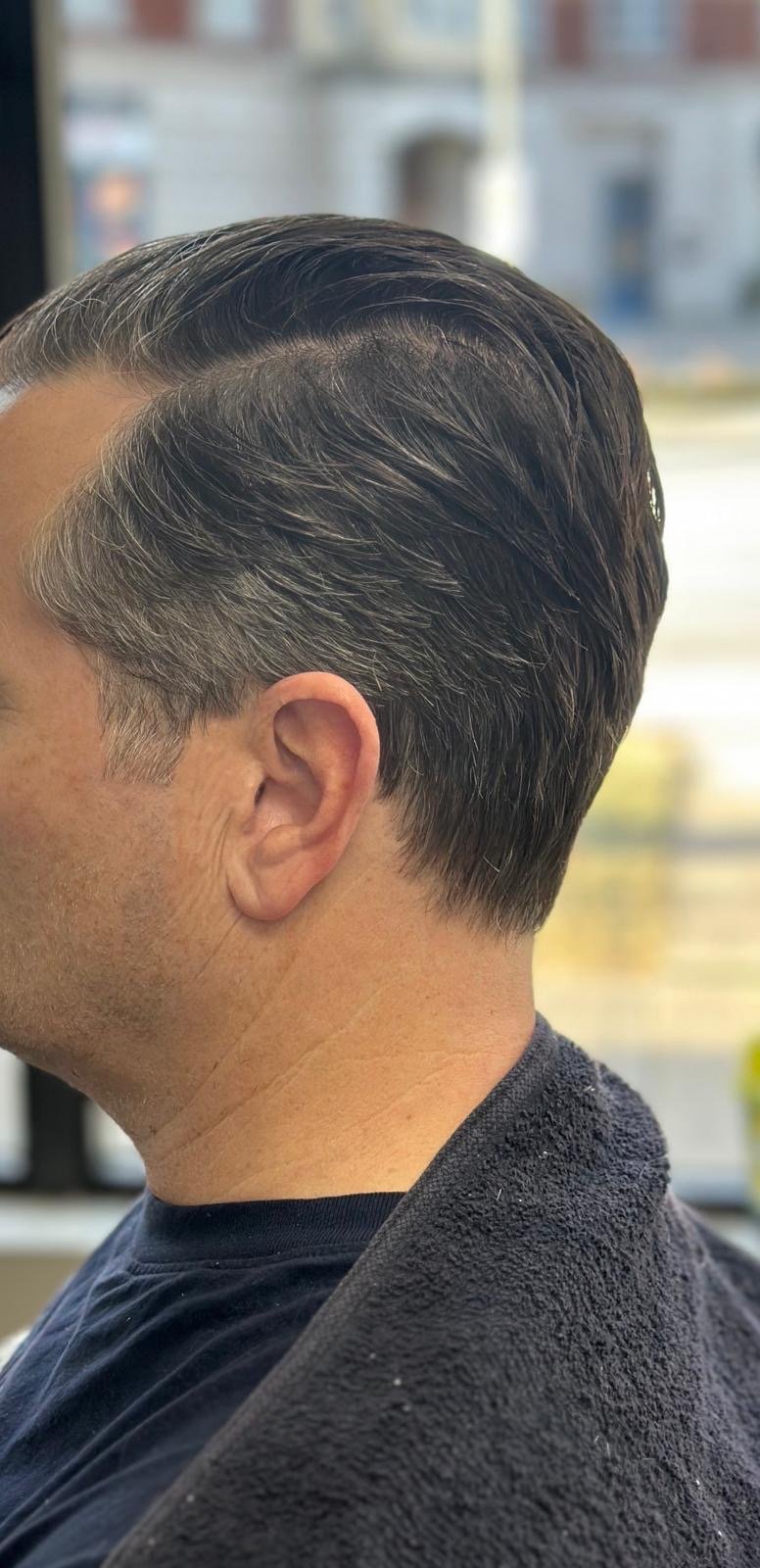 Side view of a man with dark, slicked-back hair and gray at the temples, sitting in a salon chair with a black towel around his shoulders.