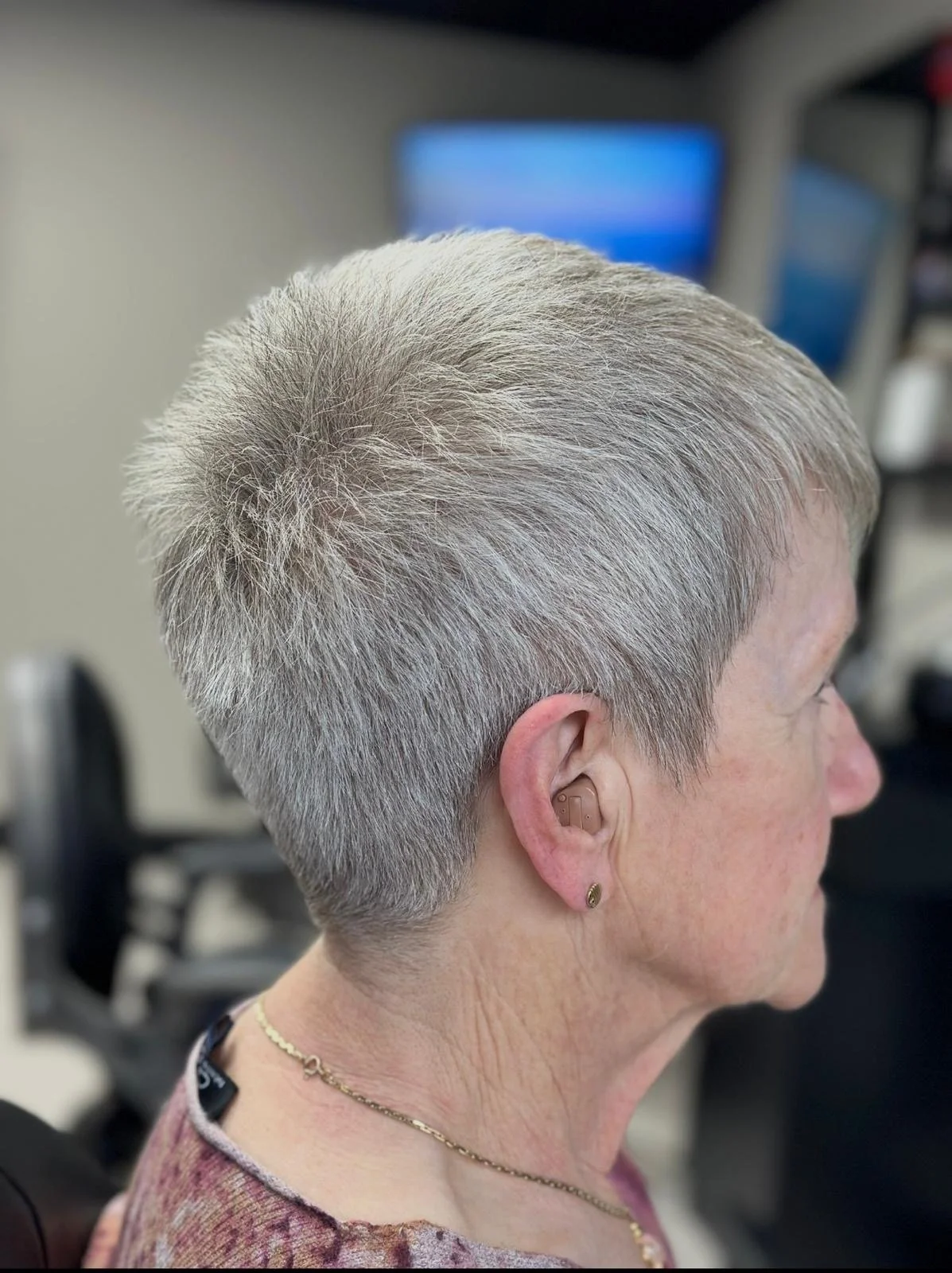 Side view of an elderly woman with short, gray, layered haircut, wearing a hearing aid and gold jewelry in a salon or medical setting.