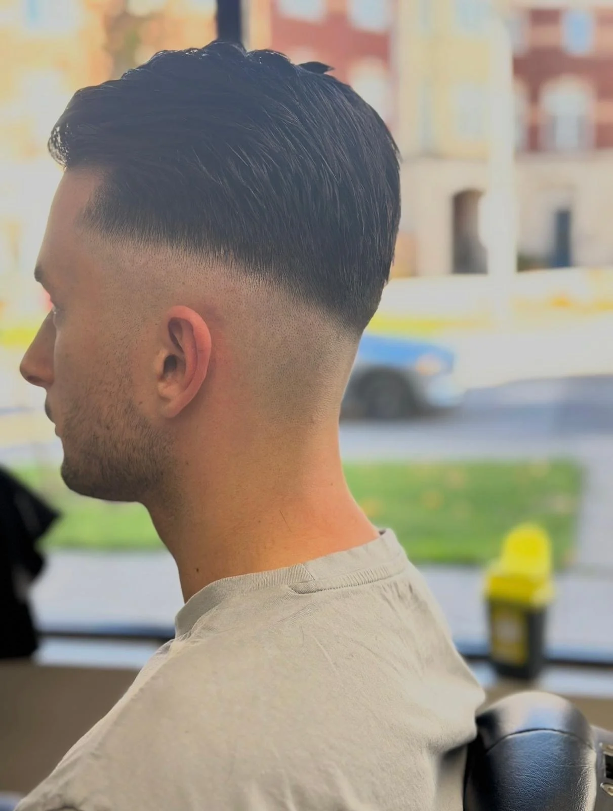 Young man with a modern fade haircut, sitting in a salon chair, with a blurred city street scene visible through the window in the background.