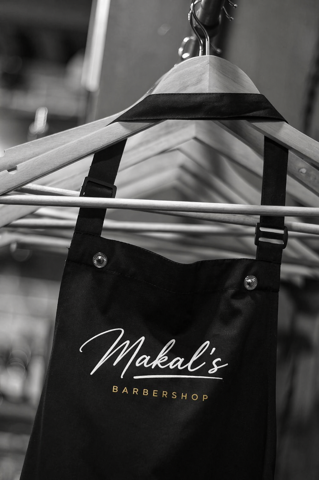 A black fabric bag with the logo 'Makali's BARBERSHOP' hanging on a wooden clothing rack with metal hangers, in a grayscale setting.