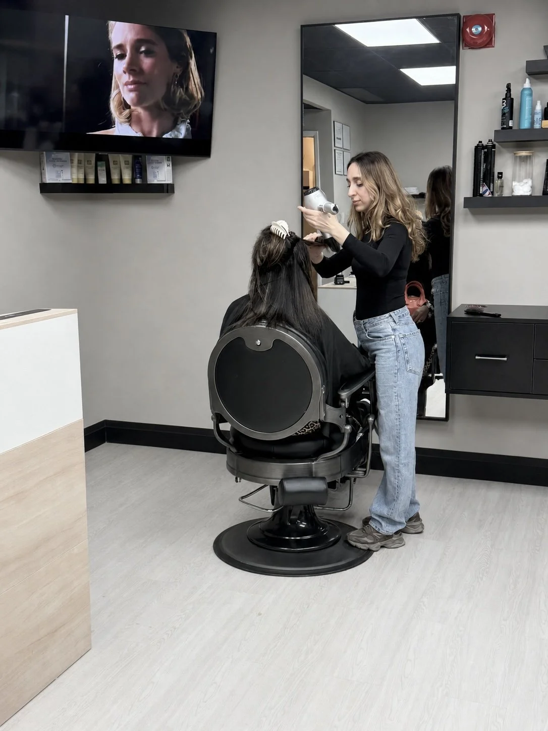 A stylist is blow-drying a woman's hair in a hair salon, with a large mirror behind them and a wall-mounted TV showing a close-up of a woman's face.