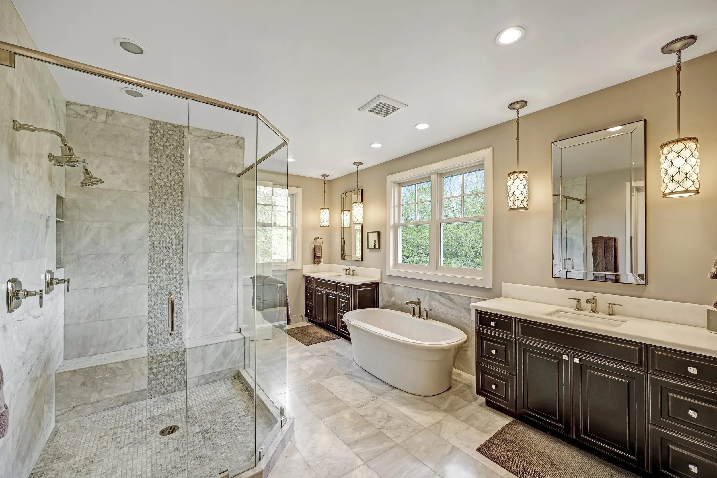 A spacious modern bathroom with a glass-enclosed shower, a freestanding bathtub, dark wood vanities, large windows, and decorative pendant lights.