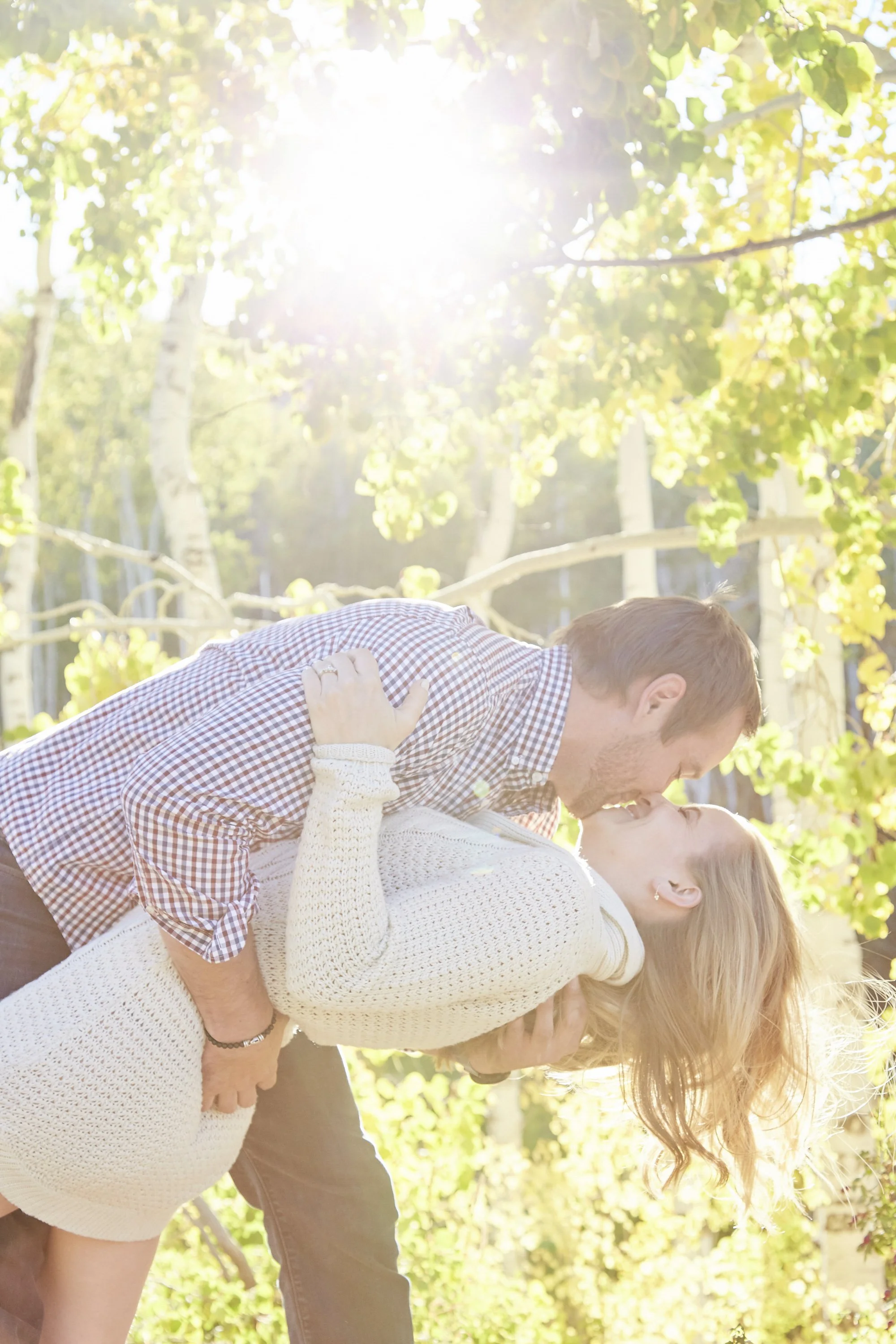 A couple is kissing outdoors in a wooded area with sunlight filtering through the trees, while a third person stands beside them