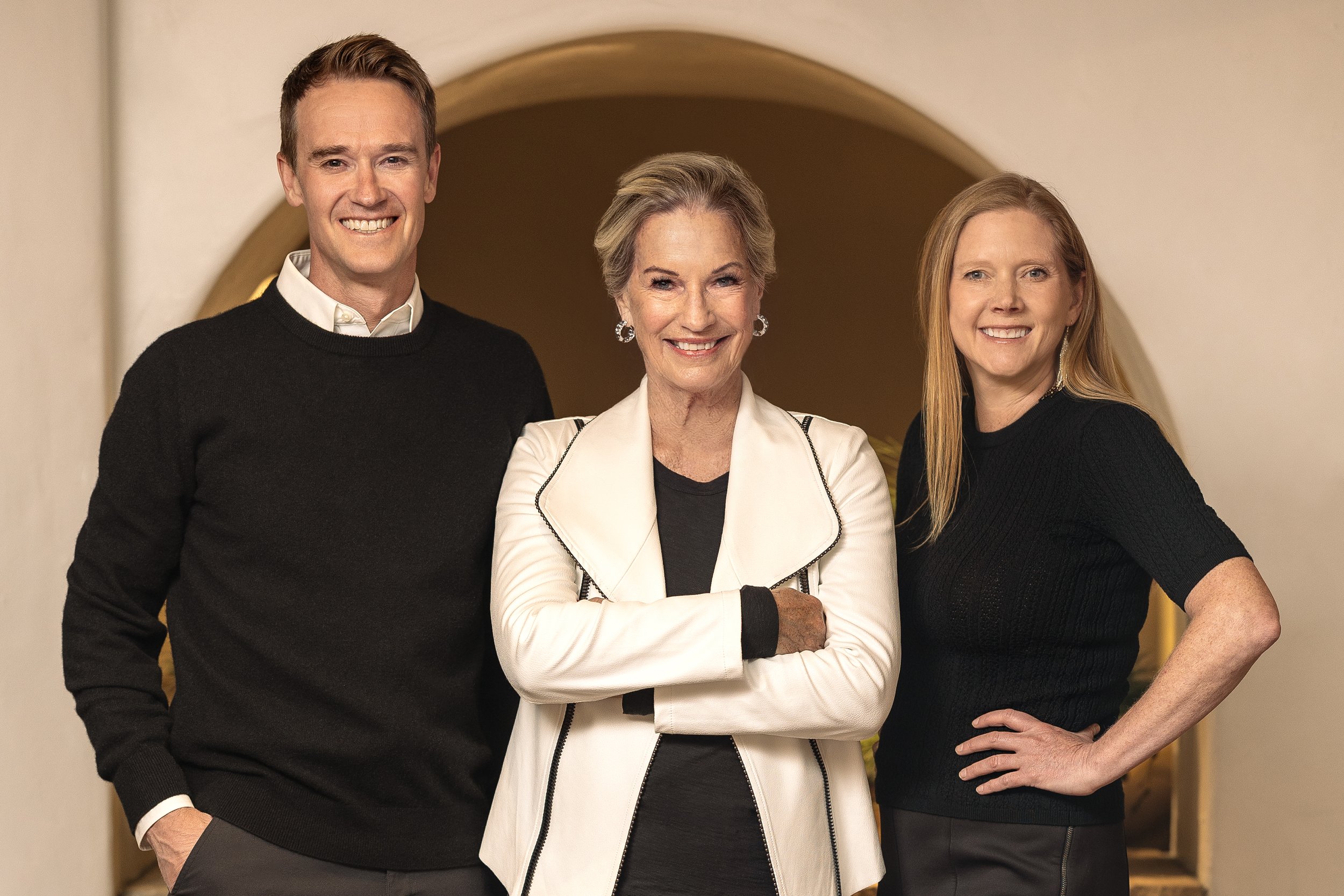 Three smiling women and one smiling man standing together indoors in front of a rounded doorway.