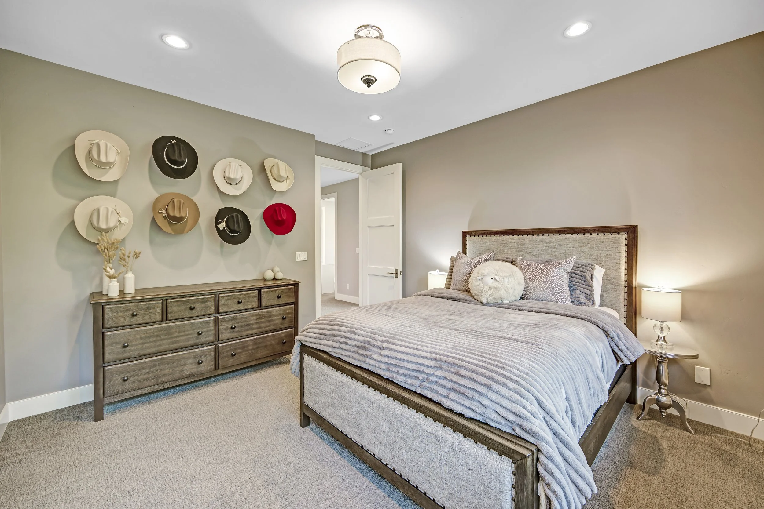A bedroom with a bed, pillows, and a headboard, a lamp on the side table, a wooden dresser, and decorative hats on the wall.