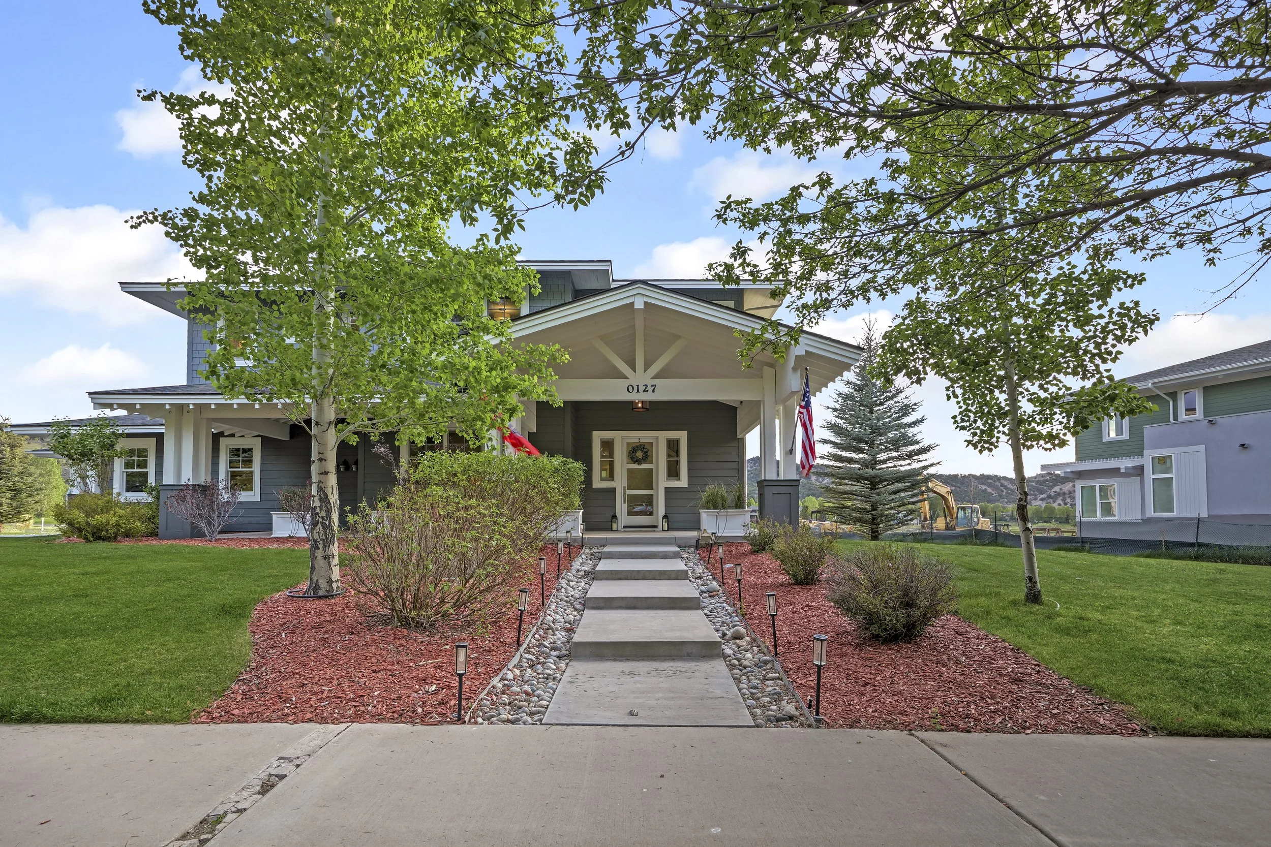 Front yard of a house with a concrete pathway leading to the front door, surrounded by red mulch, small bushes, and young trees; American flag and a wreath hang on the house; neighboring houses visible in the background; clear blue sky.