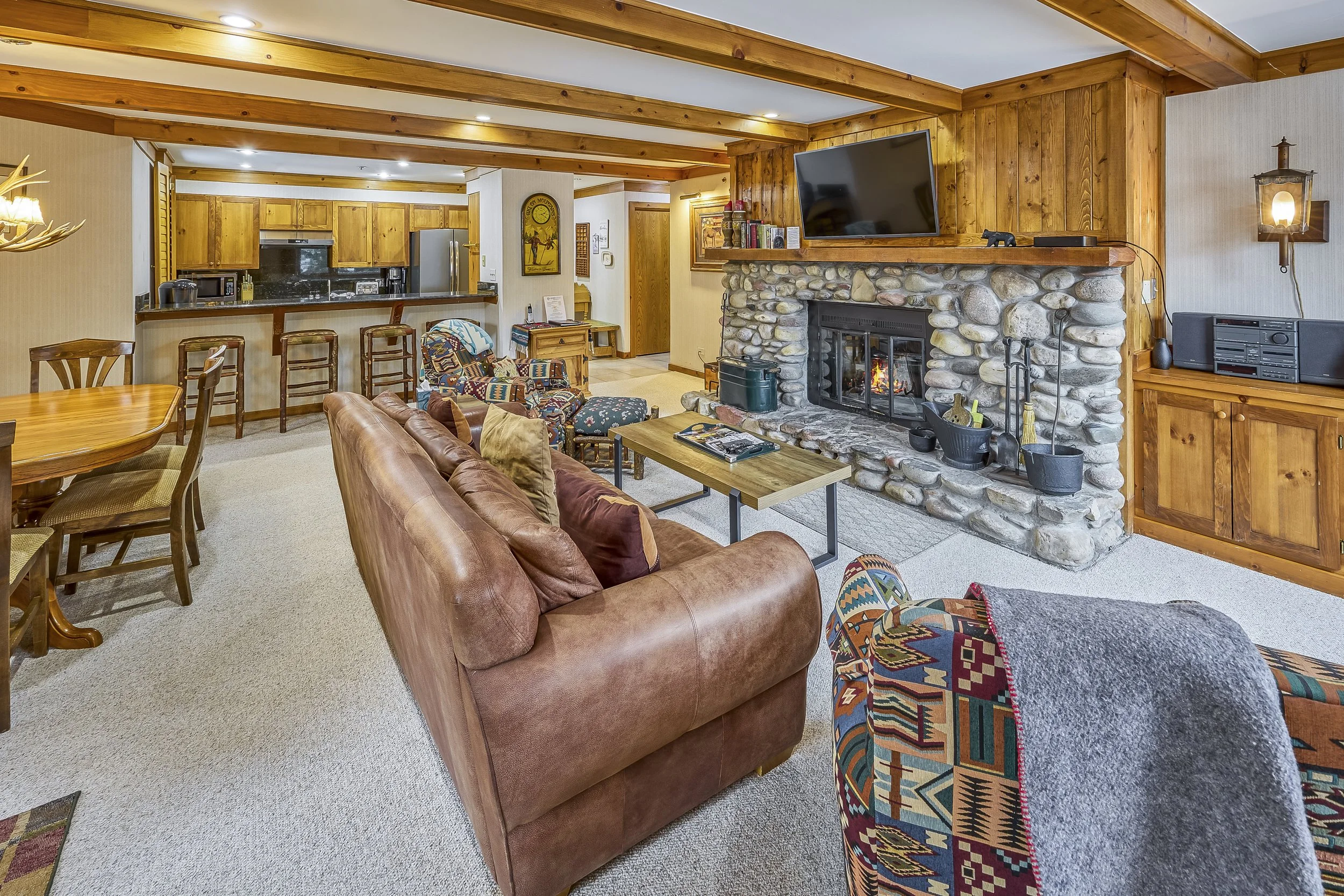 Living room with wood beams, stone fireplace, leather couch, patterned armchair, TV, and dining area in the background.