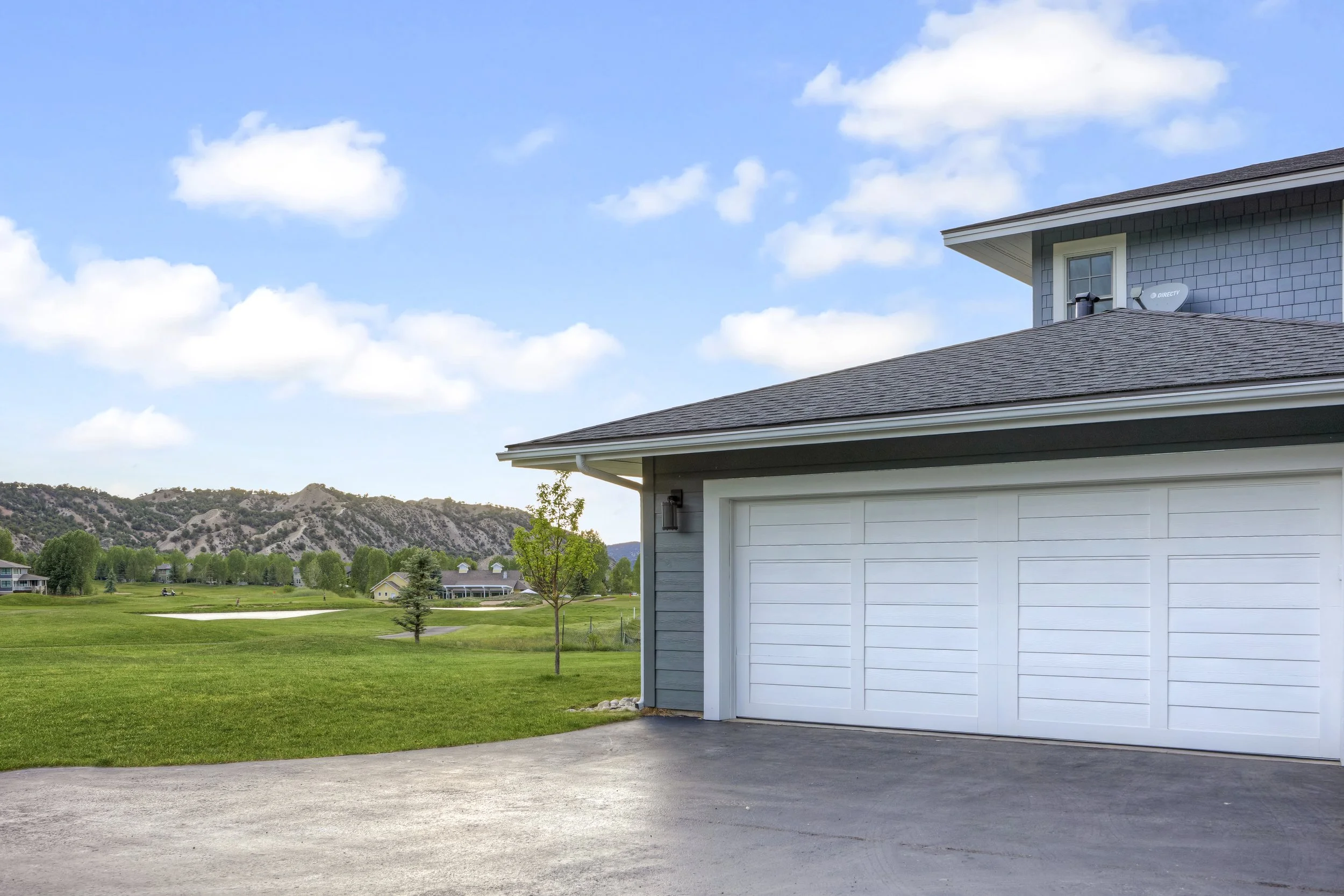 View of a house with a white garage door and blue siding, overlooking a green golf course with trees and hills in the background, under a partly cloudy sky.