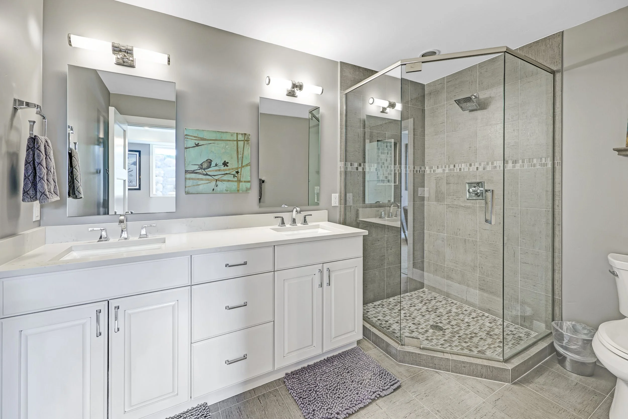 Modern bathroom with white vanity, two mirrors, gray tiled shower with glass door, and toilet, light gray walls, framed bird artwork, and gray bath mats.