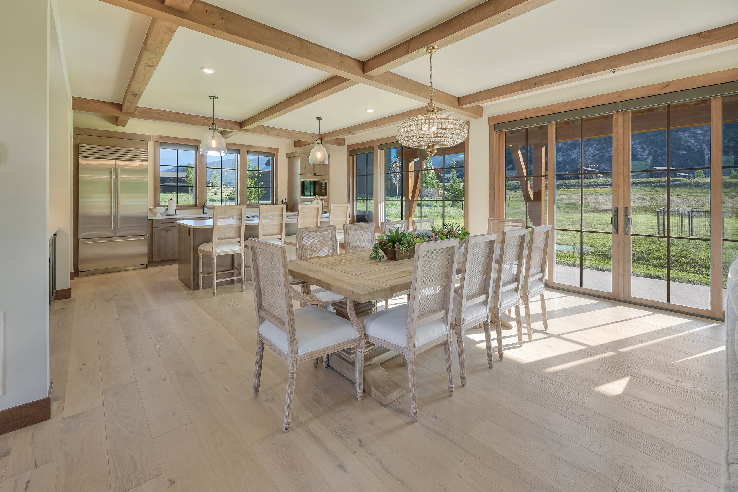 A bright, spacious kitchen and dining area with large windows overlooking a green outdoor landscape. The room features a wooden dining table with eight chairs, a chandelier, pendant lights, a stainless steel refrigerator, and a kitchen island with ba