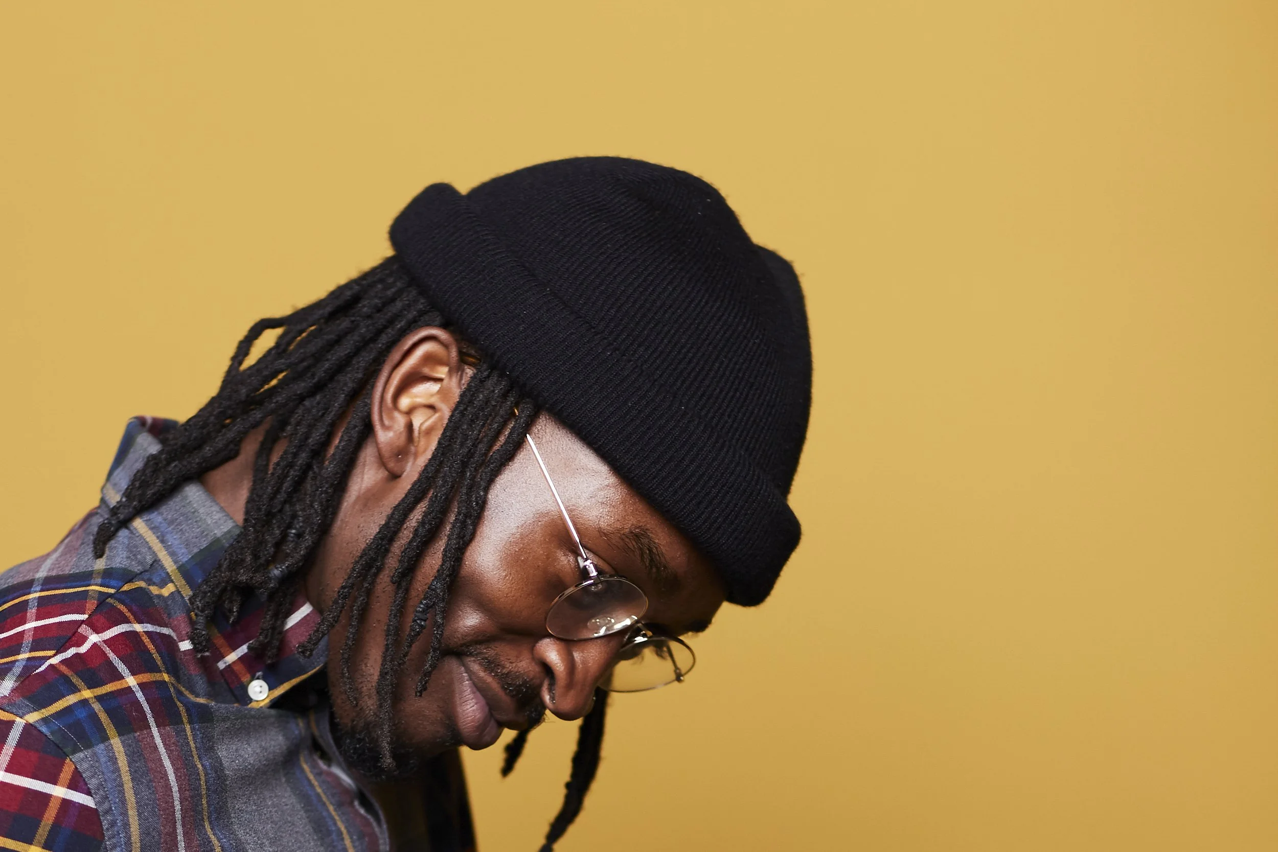 A person with dark skin, wearing glasses, a black beanie, plaid shirt, and earrings, appears to be looking downward against a solid yellow background.
