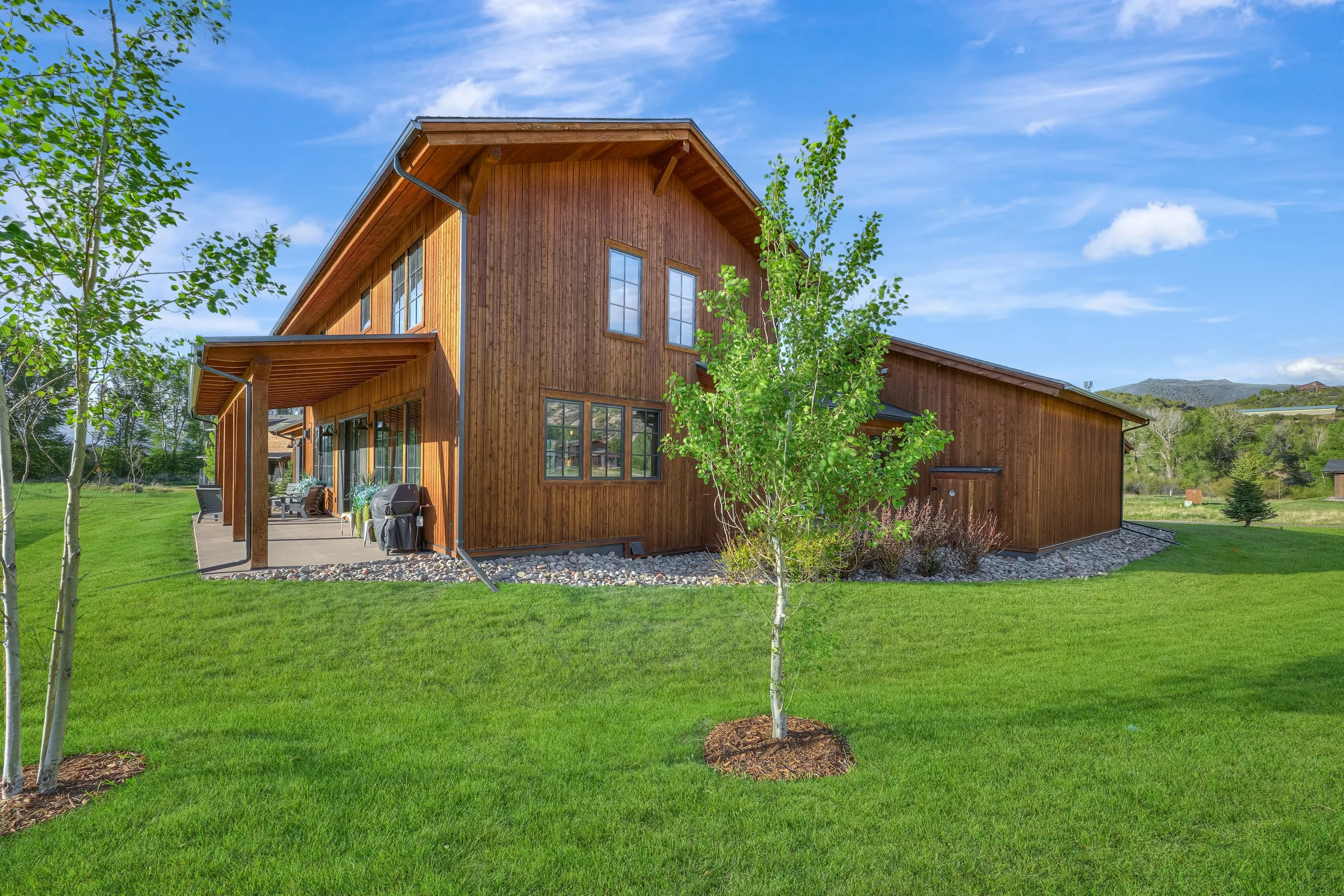 A two-story wooden house with a large patio area, surrounded by a well-manicured lawn and young trees, with mountains in the background under a blue sky with some clouds.