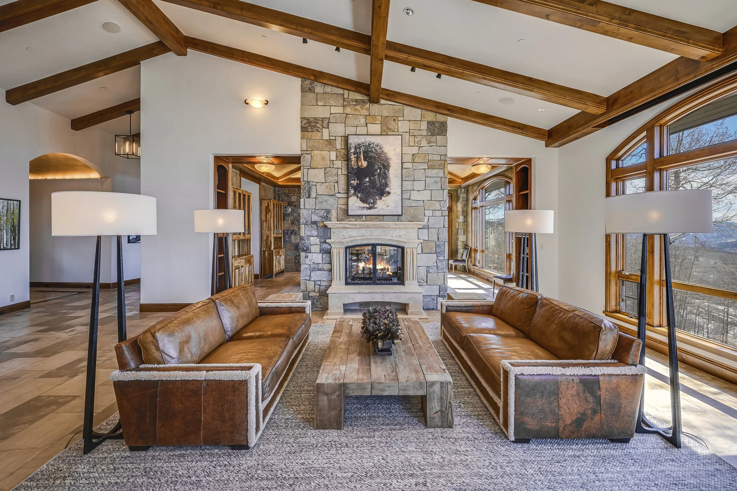 Living room with leather sofas, stone fireplace, large windows, and wooden ceiling beams.