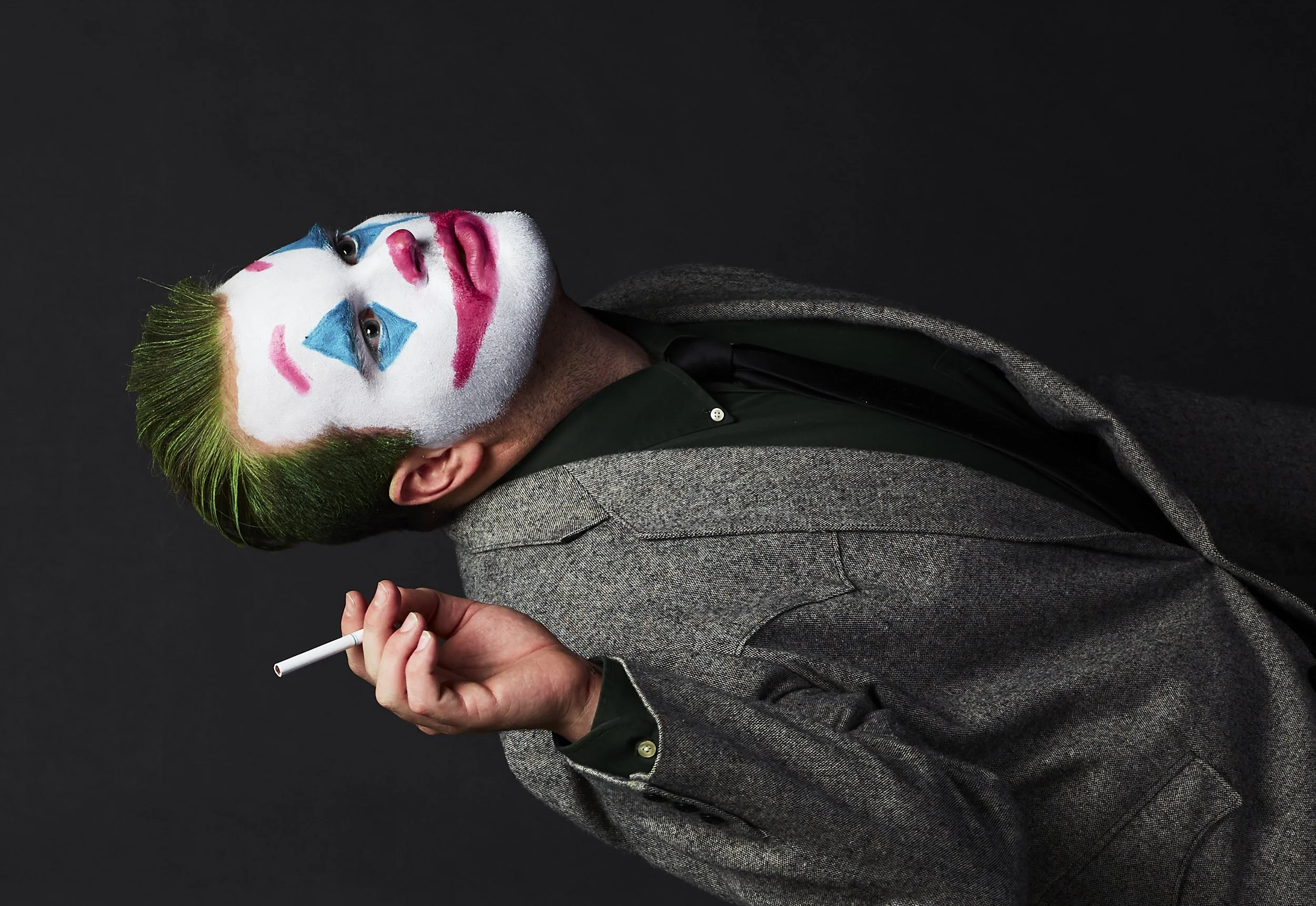 A person with clown makeup and green hair, wearing a gray blazer and a dark shirt, holding a cigarette in one hand, against a dark background.