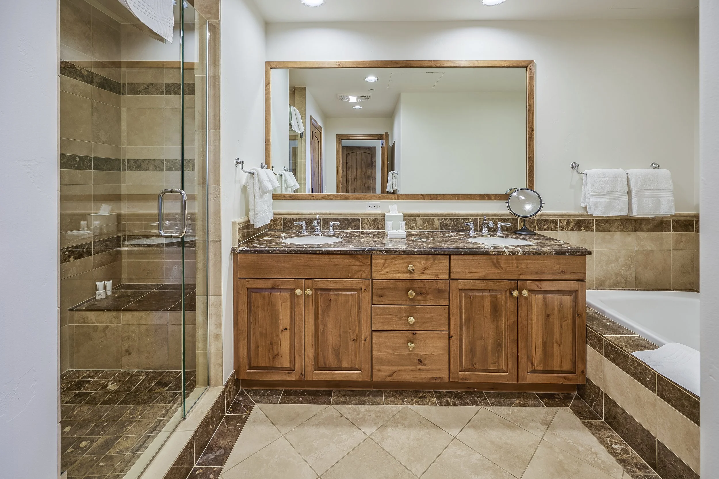 A bathroom with a large mirror above a wooden vanity with two sinks and a marble countertop, a glass-enclosed shower on the left, and a bathtub on the right. The bathroom has beige and brown tiled surfaces and white towels.