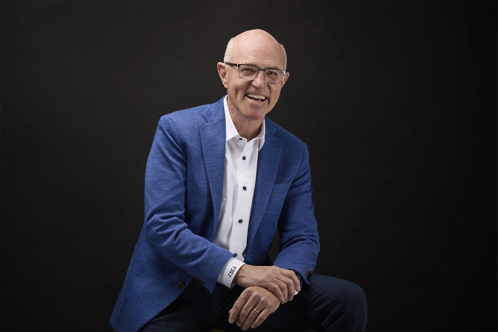 An older man with glasses, a white shirt, and a blue blazer, smiling and sitting against a plain black background.