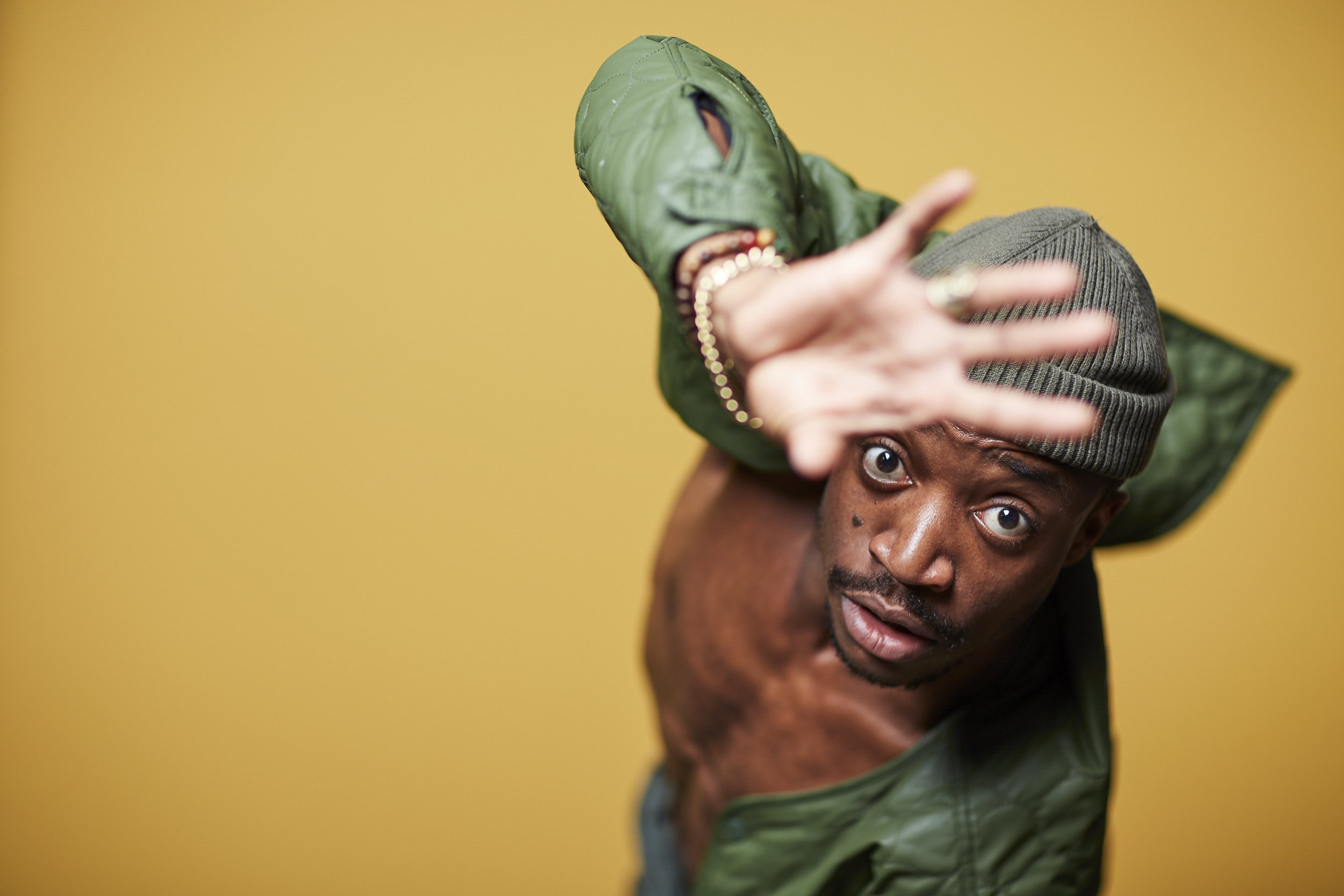 A person wearing a gray beanie and a green jacket, looking directly at the camera with wide eyes, against a yellow background, reaching out towards the camera with a blurred hand.