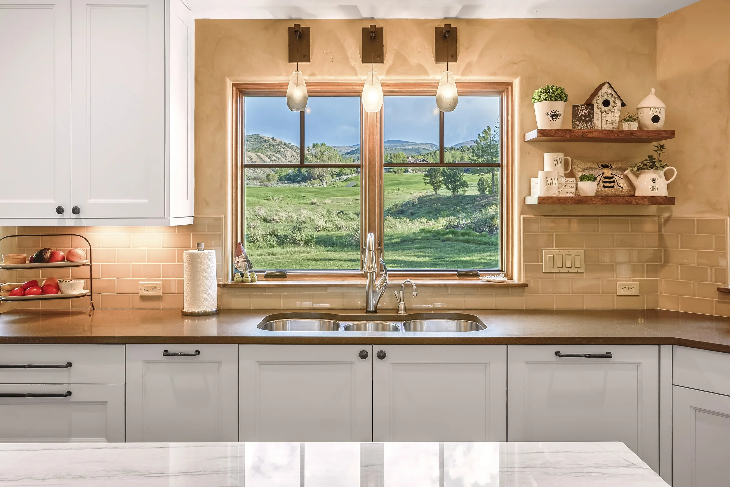 Kitchen view through window showing green grass, trees, and hills, with three pendant lights hanging above a kitchen sink, white cabinets, and a beige backsplash.