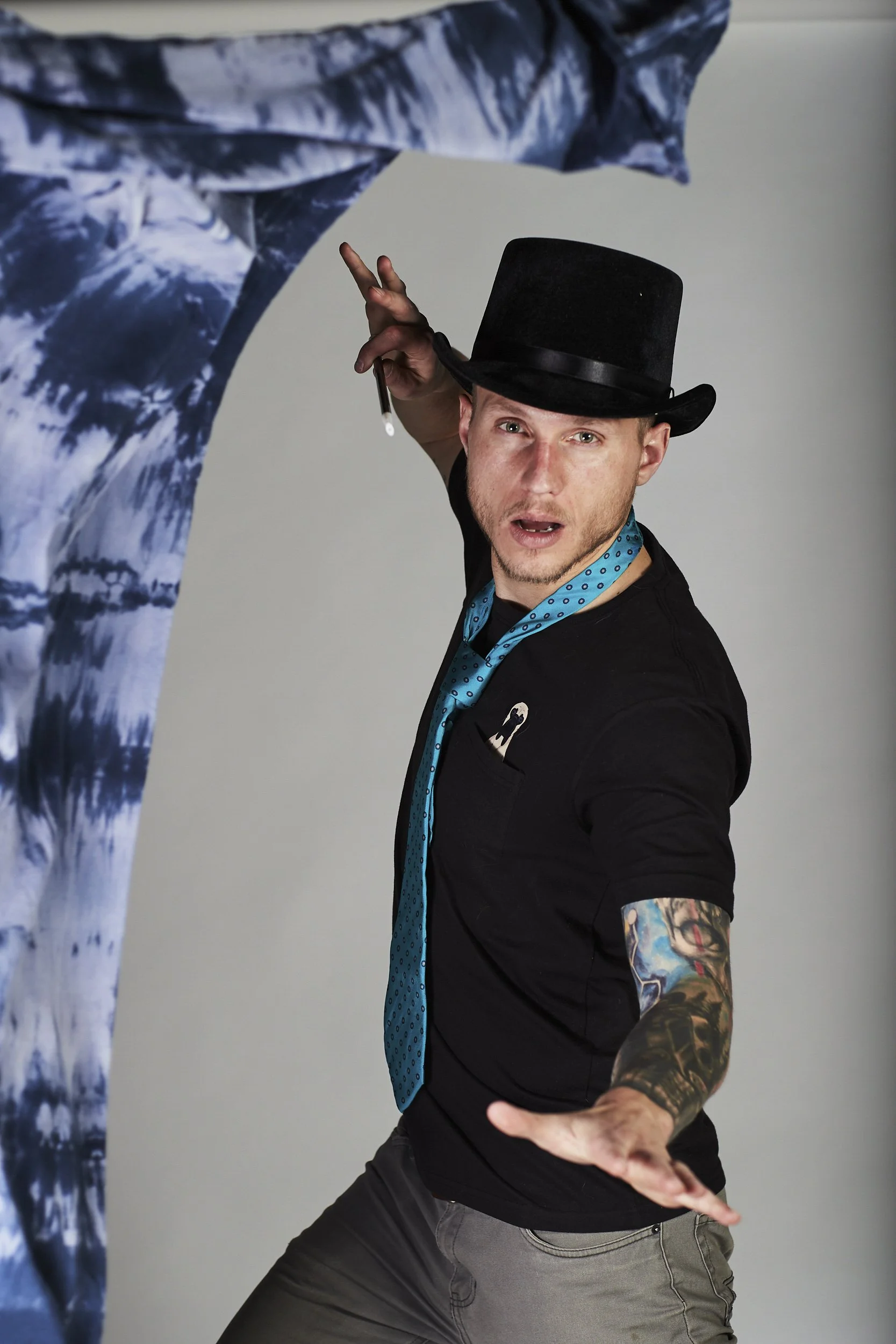 A man wearing a black top hat, black T-shirt, and gray pants, with colorful tattooed sleeves, striking a pose with one hand extended and finger pointed. A large piece of artwork or fabric with blue and white patterns is visible behind him.