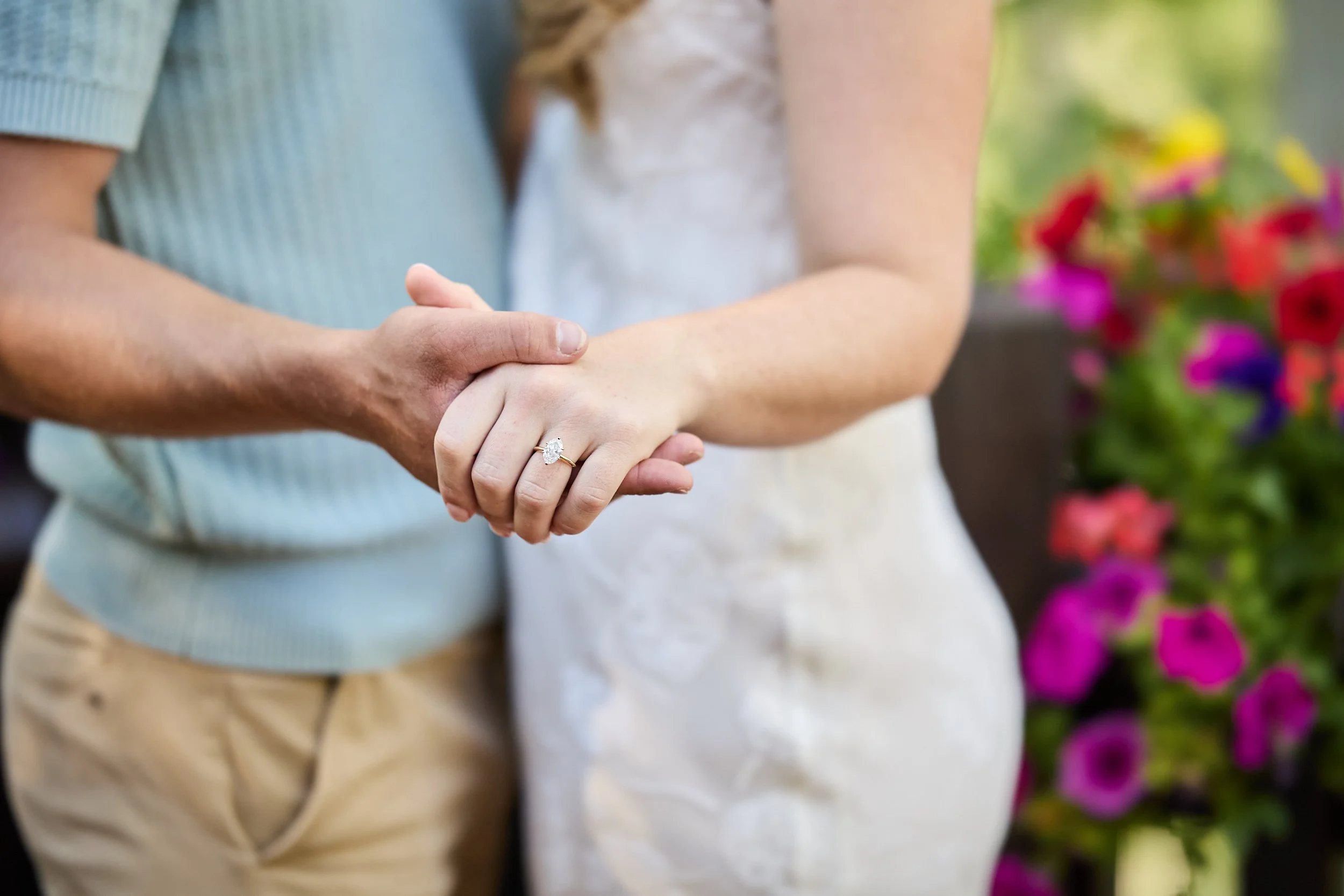 Close-up of an engaged couple holding hands, with the woman wearing an engagement ring, standing outdoors near colorful flowers.
