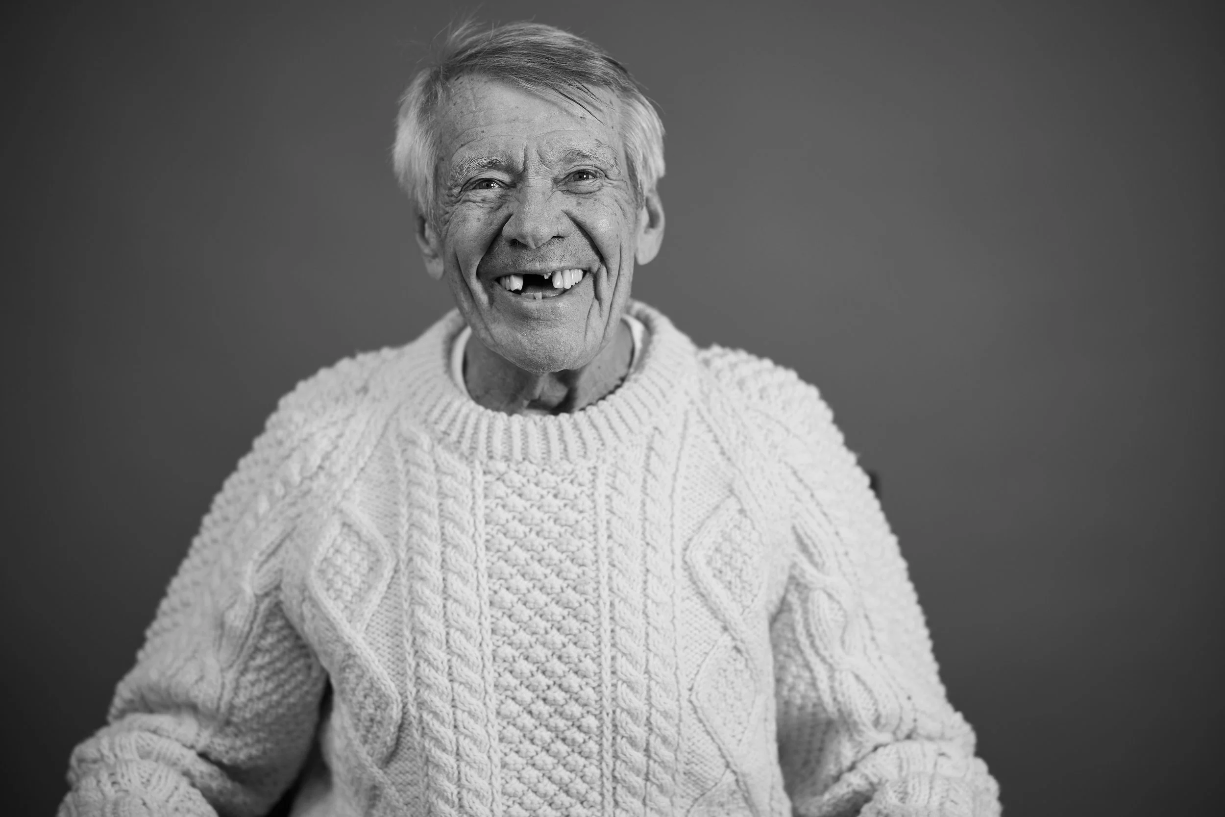 An elderly man with gray hair, missing front teeth, and wrinkles, smiling broadly. He wears a chunky knit sweater and stands against a plain, dark background.