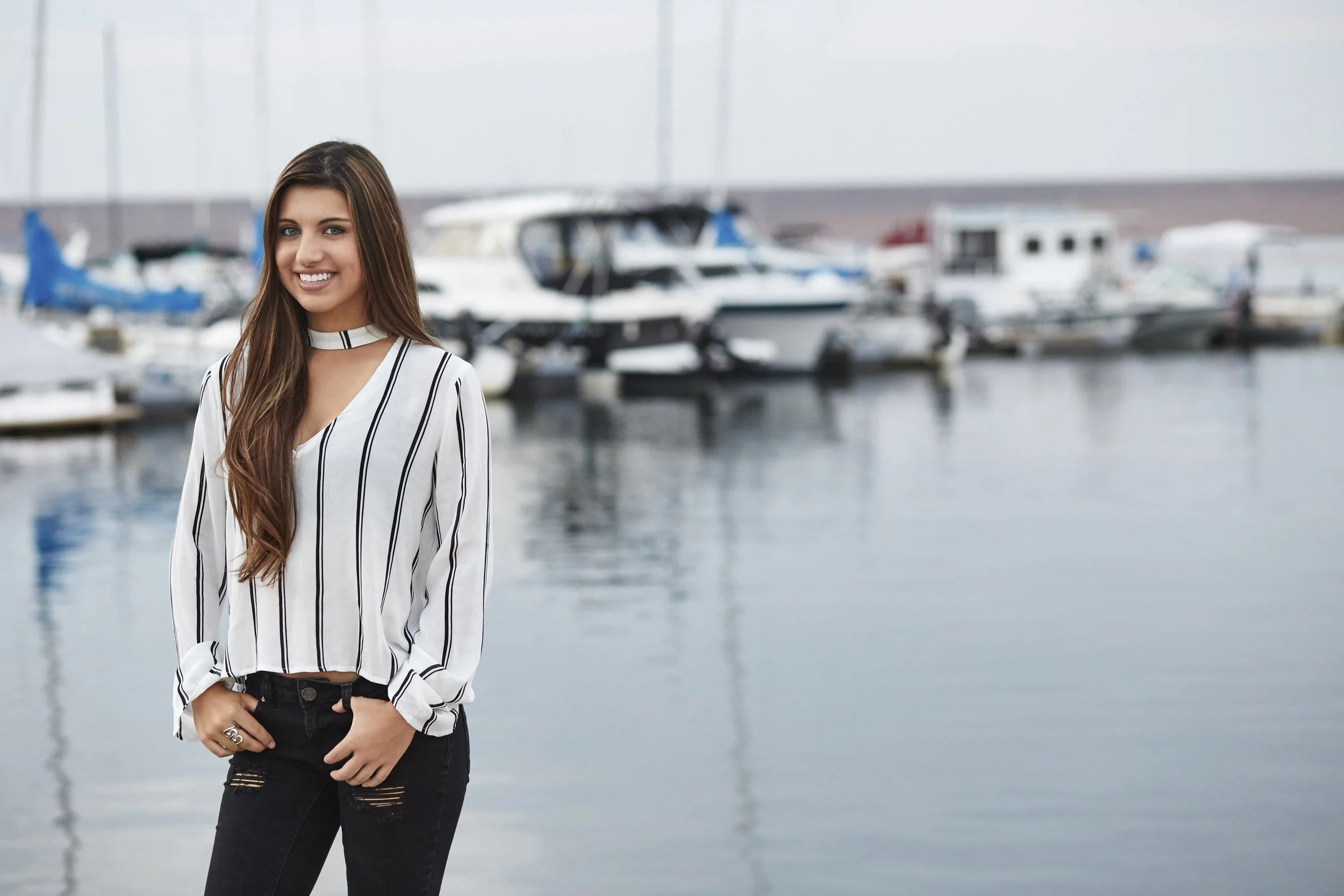 A young woman with long brown hair smiling at the camera, standing at a marina with boats docked in the background.