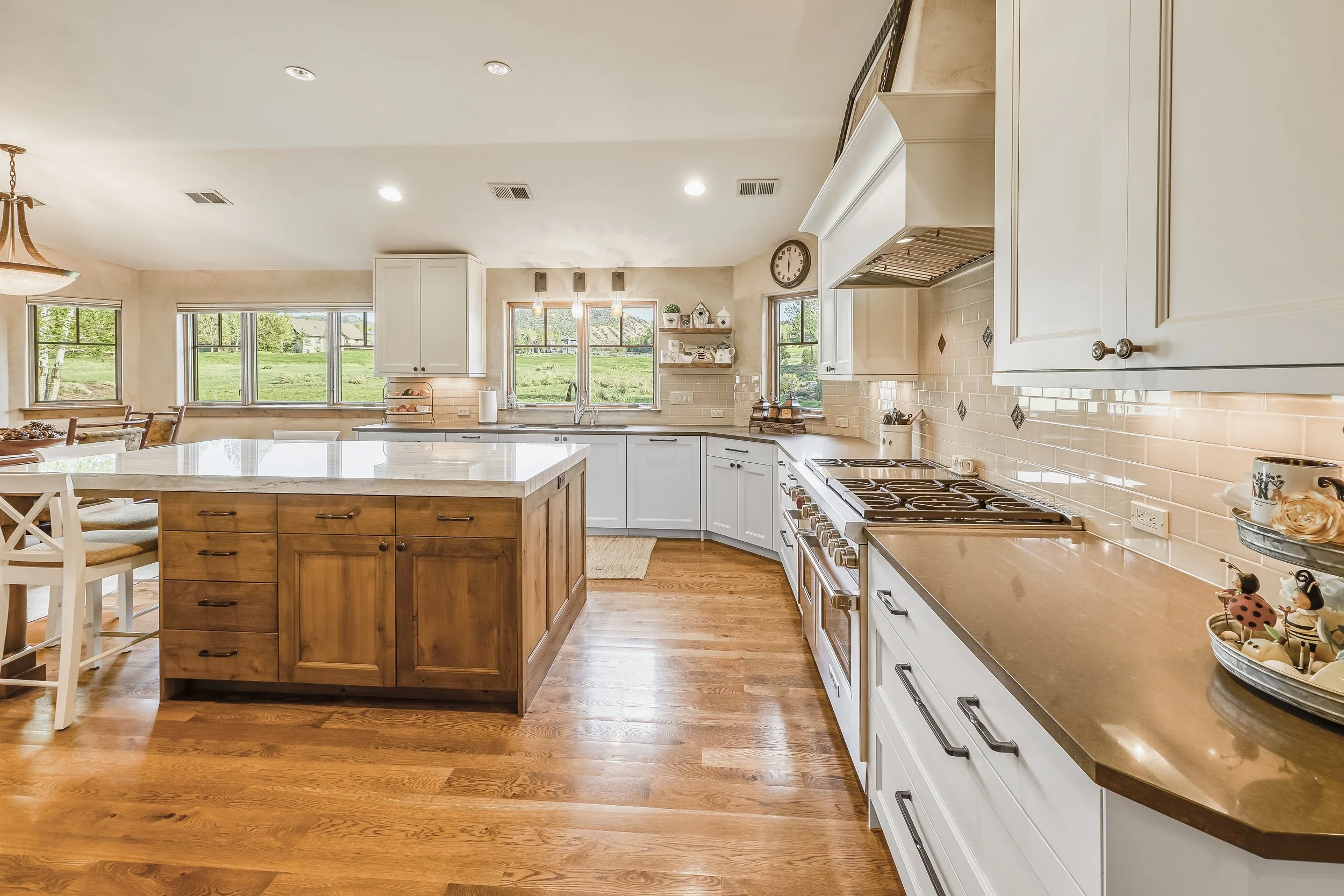 Bright kitchen with white cabinets, a large island with a wooden base, and hardwood floors. There are large windows overlooking a green lawn. Kitchen appliances include a stove, and decorative items are on the counters and open shelves. Recessed and 