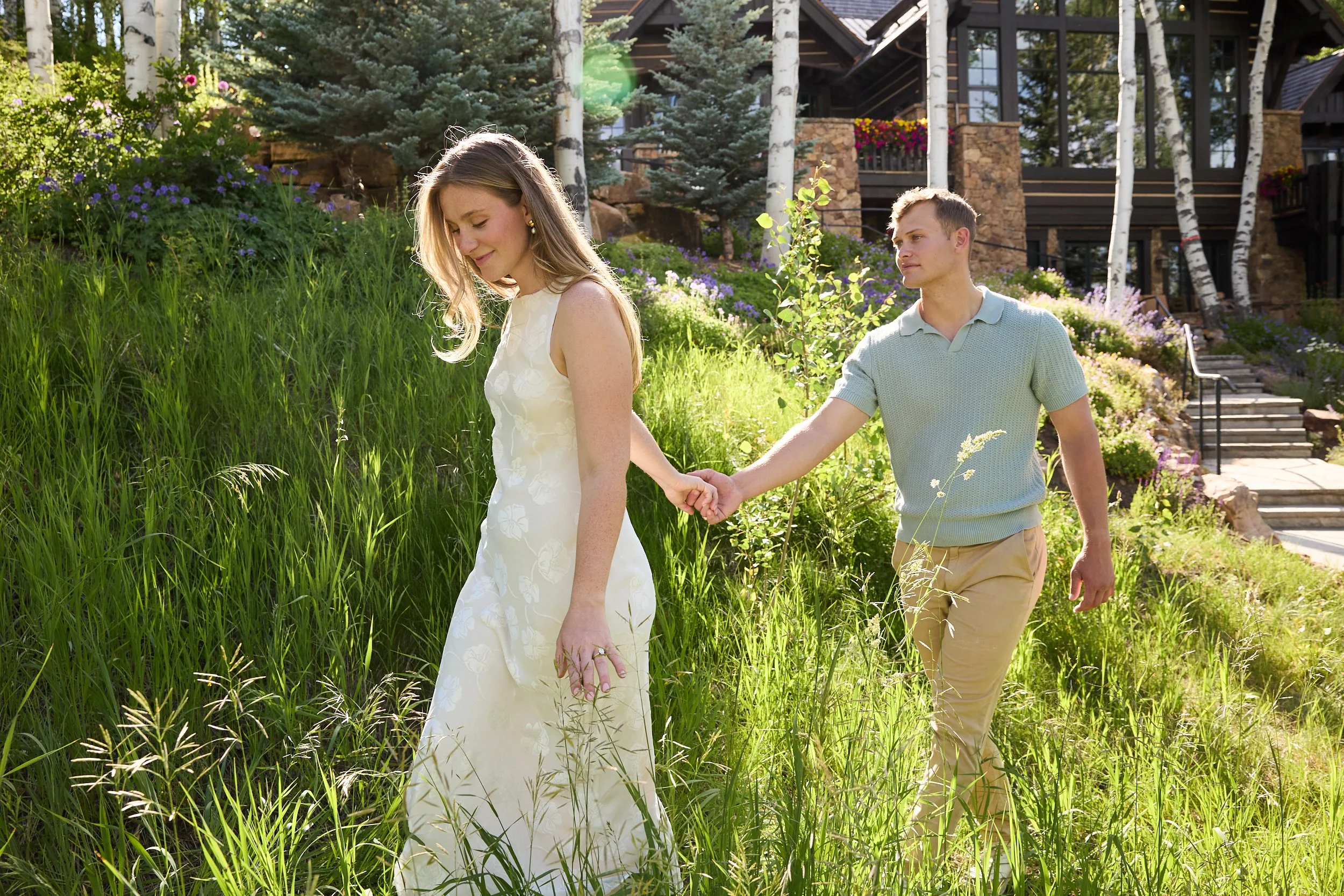 A young woman in a white dress walks through tall grass, holding hands with a young man in a light blue polo shirt on a sunny day in a garden with trees and a house in the background.