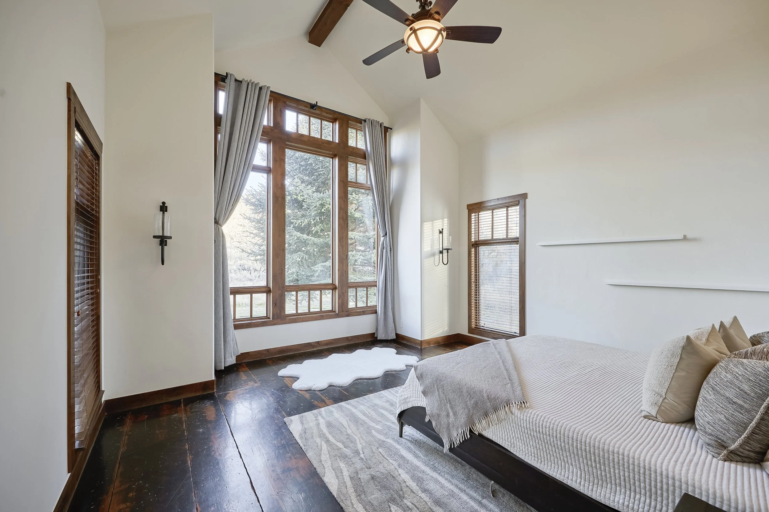 A bright living room with large wooden framed windows, curtains, a ceiling fan, and a bed with pillows.