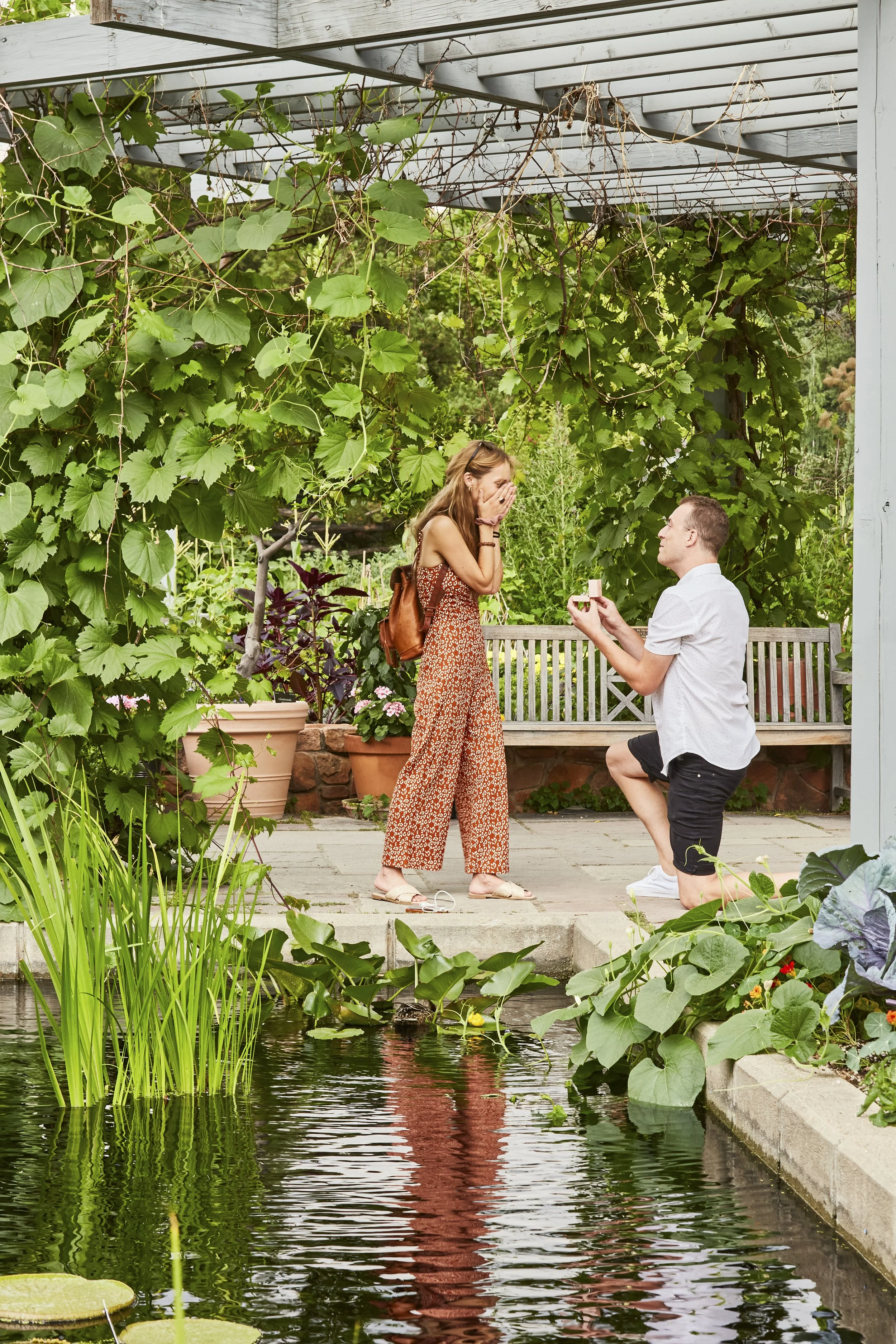 A man proposing to a woman by a pond in a lush garden setting. The man is kneeling on one knee holding a small box, and the woman looks surprised and happy, covering her mouth with her hands.