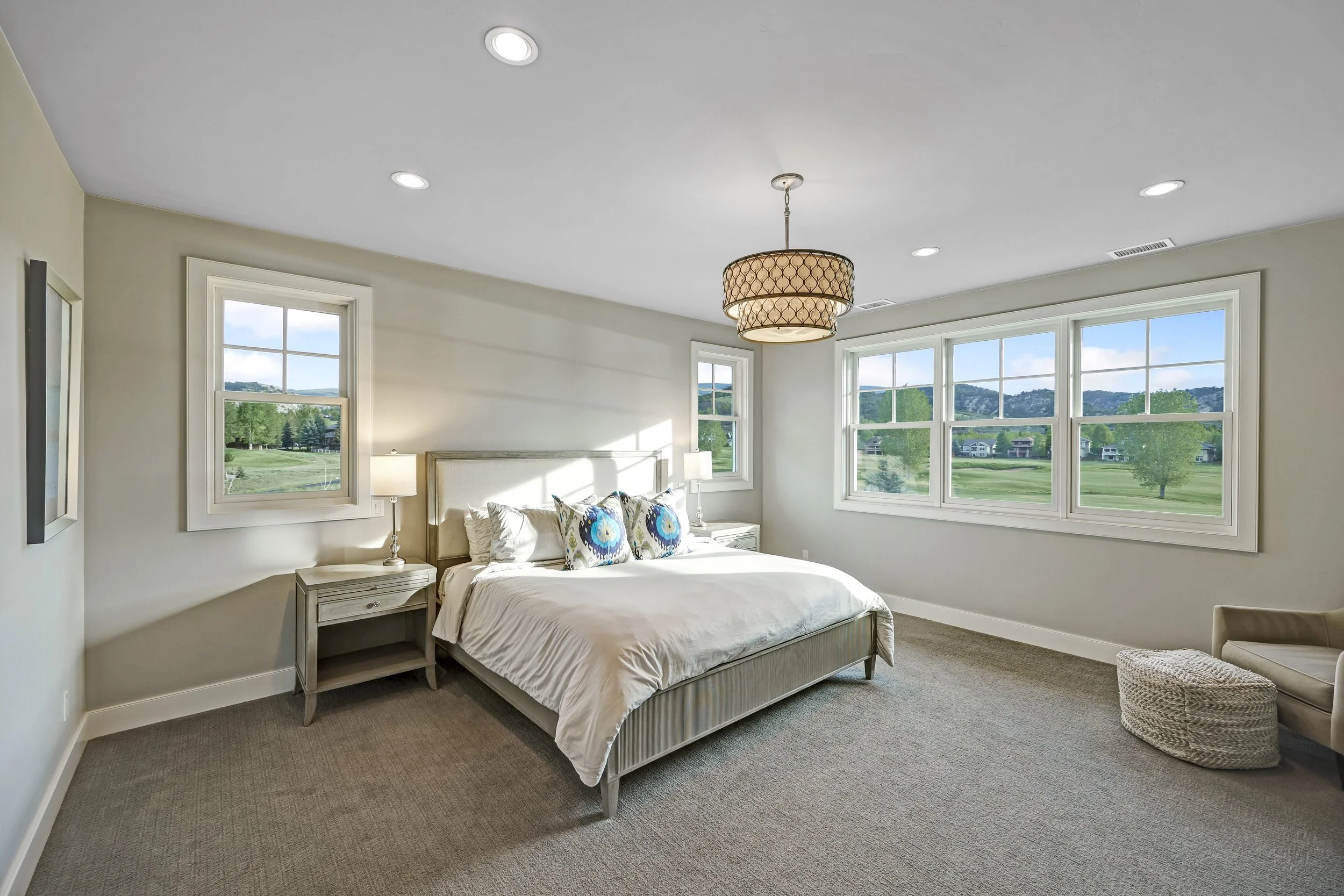 Bright bedroom with large windows overlooking green landscape, white bed with blue and white patterned pillows, bedside lamps, and beige carpet.