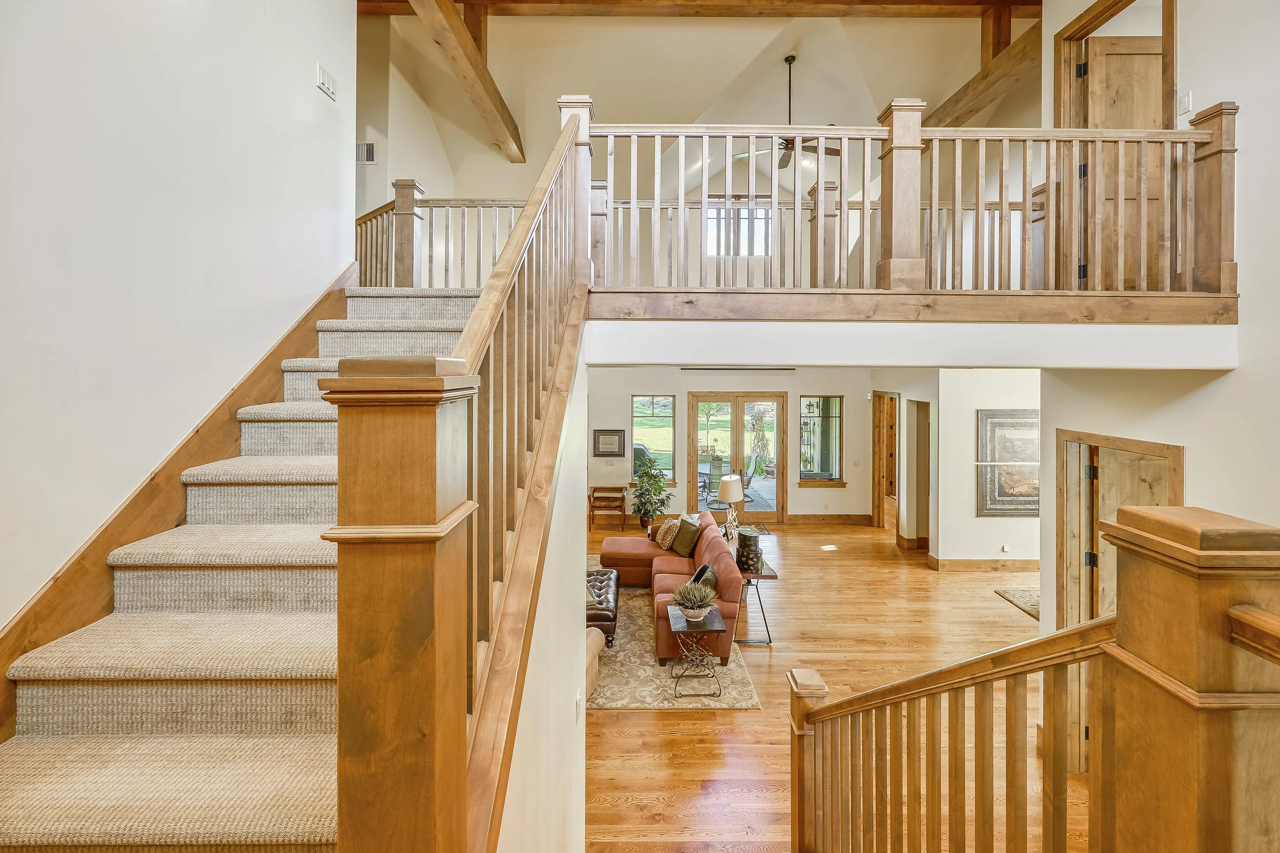 Interior view of a two-story home with wooden staircase, railing, and beams, showcasing a living room with a sofa, chairs, and large windows allowing natural light.