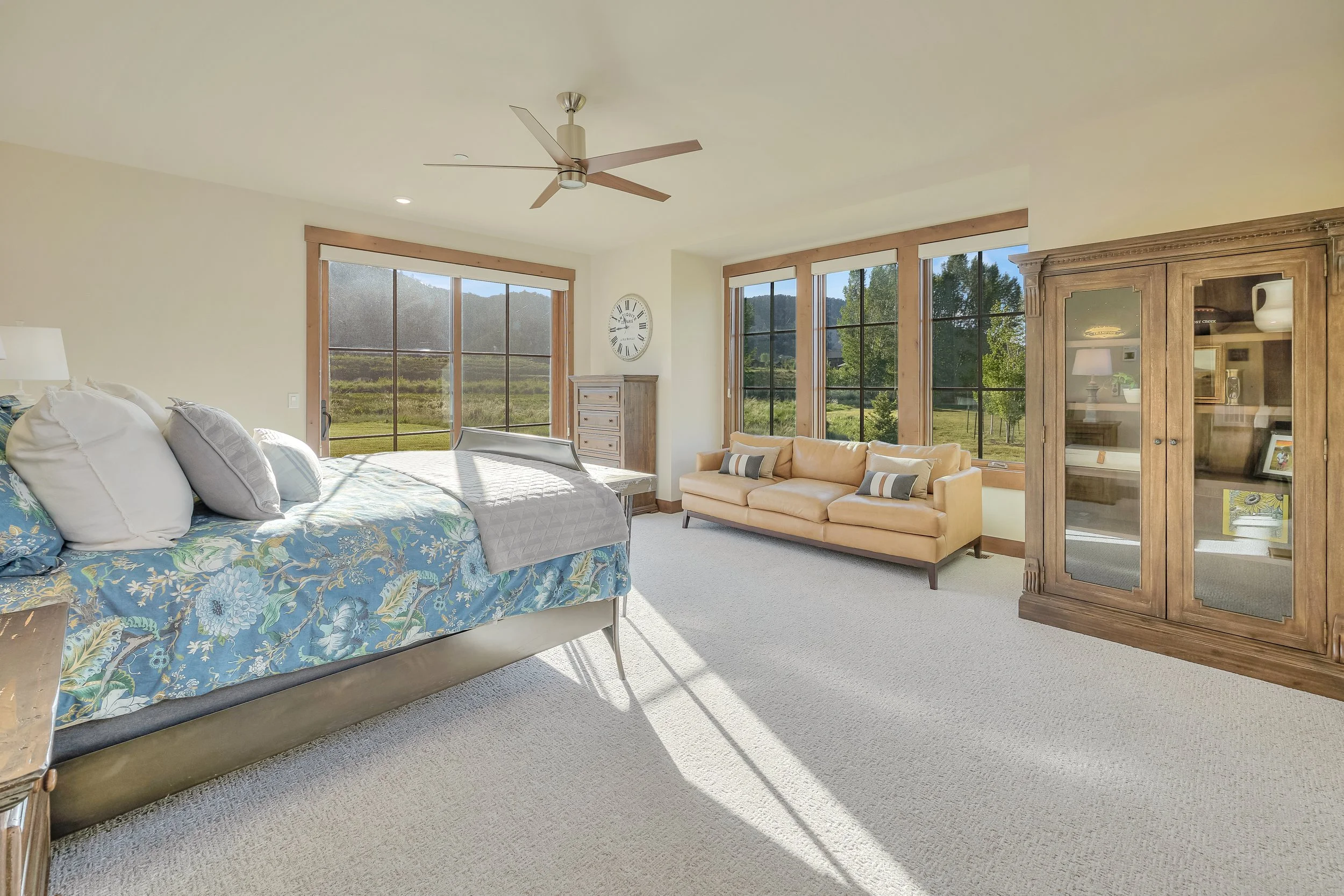 A spacious bedroom with large windows showcasing a scenic outdoor view, a bed with a floral blue bedspread and pillows, a wooden dresser, a beige sofa with pillows, an antique wooden cabinet, and a ceiling fan.