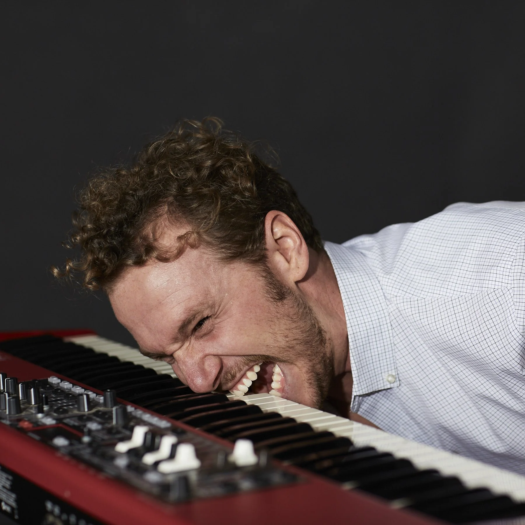 A young man with curly hair and a beard is playing a keyboard with joy, leaning over and smiling as he presses the keys.