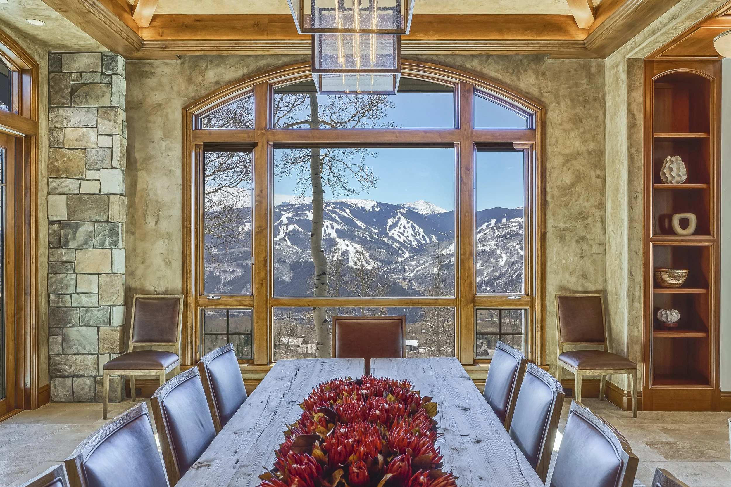 The image shows a modern dining room with a large window overlooking snow-capped mountains and a leafless tree. The room has a wooden ceiling with a rectangular chandelier hanging above a long wooden dining table. There are chairs around the table, and a decorative centerpiece of red flowers in the middle of the table.
