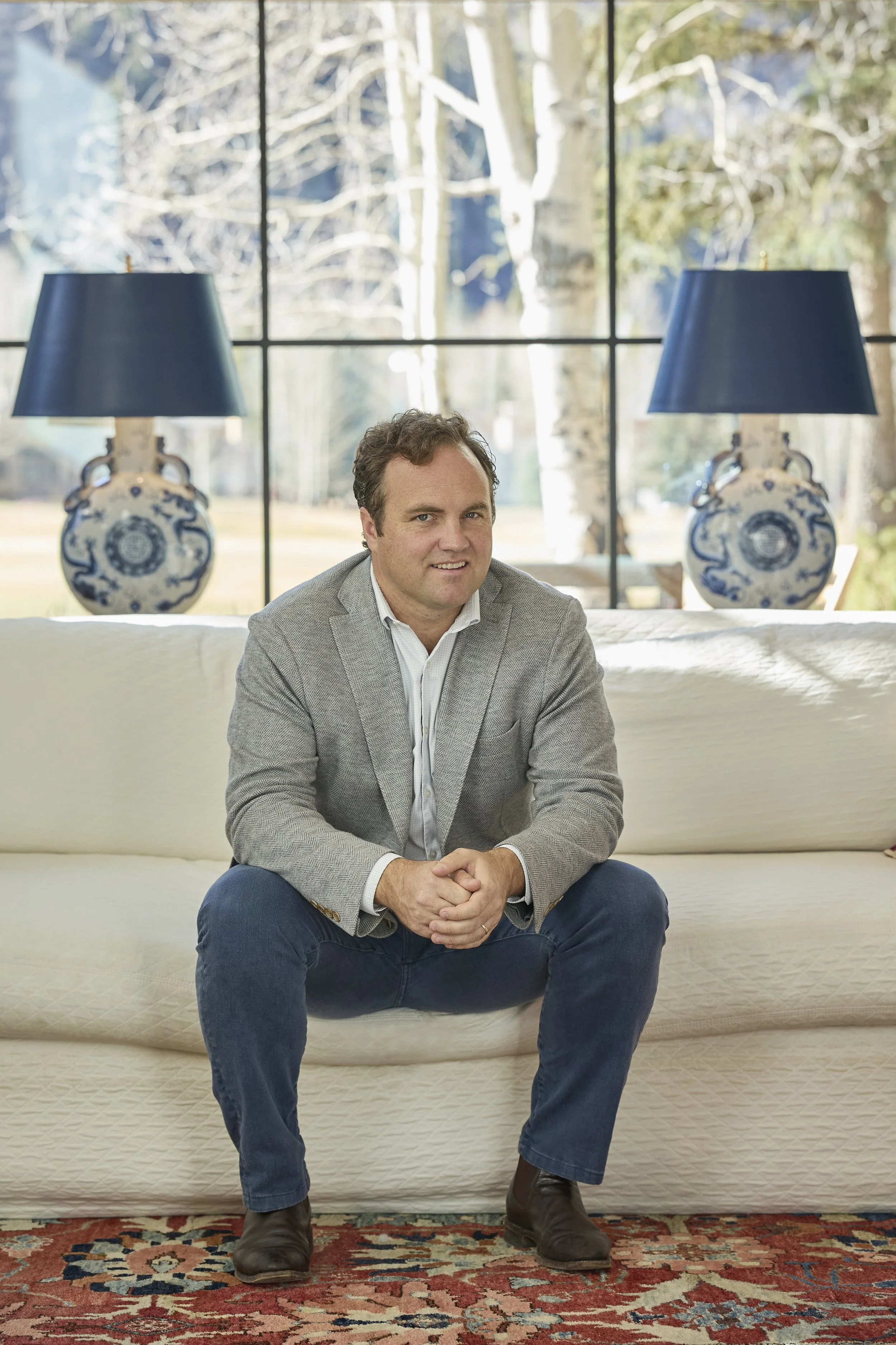A man sitting on a cream-colored sofa in a room with large windows and a view of trees outside. He is wearing a gray blazer, white shirt, blue jeans, and brown boots. Two blue lamps with white and blue decorative bases are on either side of the windo