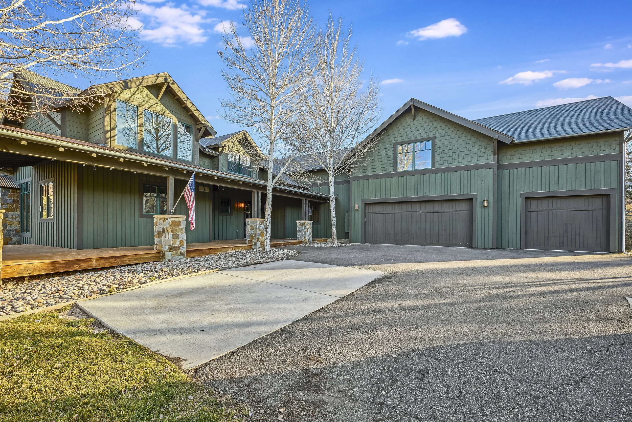 View of a modern two-story house with green siding, large windows, stone accents, a wooden deck with an American flag, a double garage, leafless trees, and a driveway with a gravel strip, under a blue sky with some clouds.