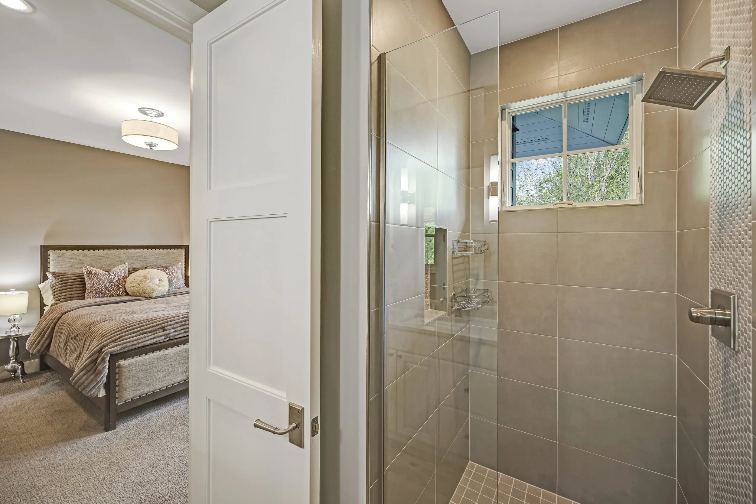 A bathroom with beige tiled walls and a small window showing trees outside. There is a large overhead shower with a square showerhead and built-in shelves, and a vanity with boxes on a shelf that match the bathroom decor.