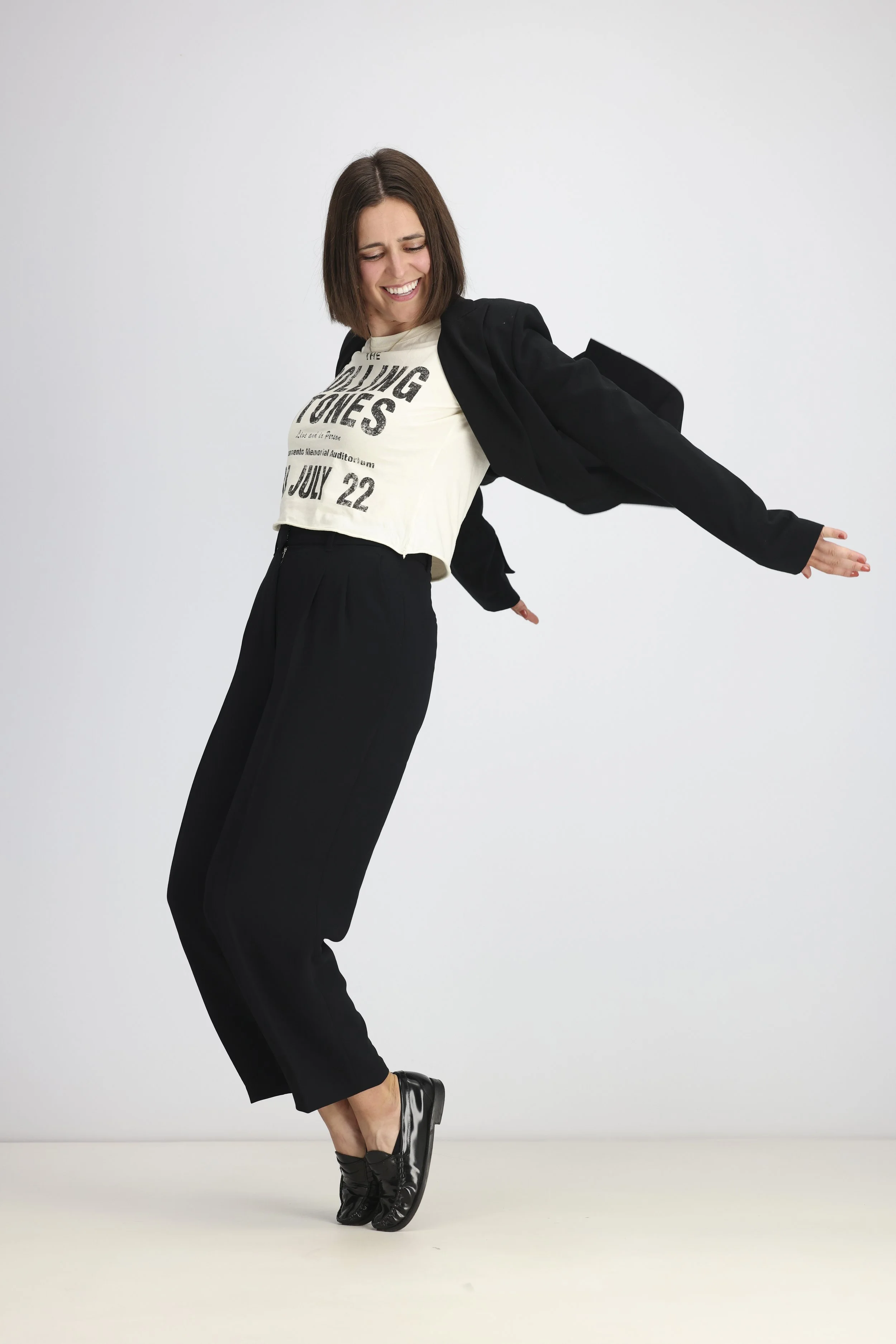 A woman with short brown hair wearing a black blazer, loose black pants, a cream graphic t-shirt, and shiny black shoes. She is standing on one leg with arms outstretched and a joyful expression.