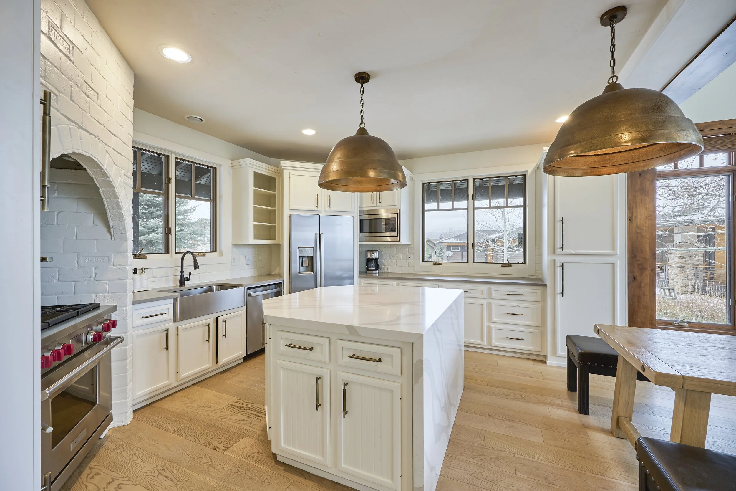 Bright kitchen with white cabinets, stainless steel appliances, a kitchen island, light wood flooring, large windows, and hanging brass pendant lights.
