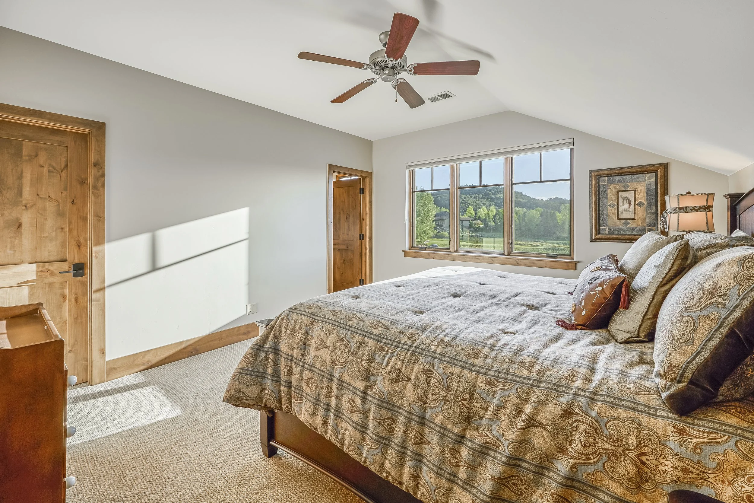 A bedroom with a large bed covered in a paisley patterned bedspread, pillows, and a window with a scenic view of greenery and hills outside, a ceiling fan with wooden blades, and framed wall art.