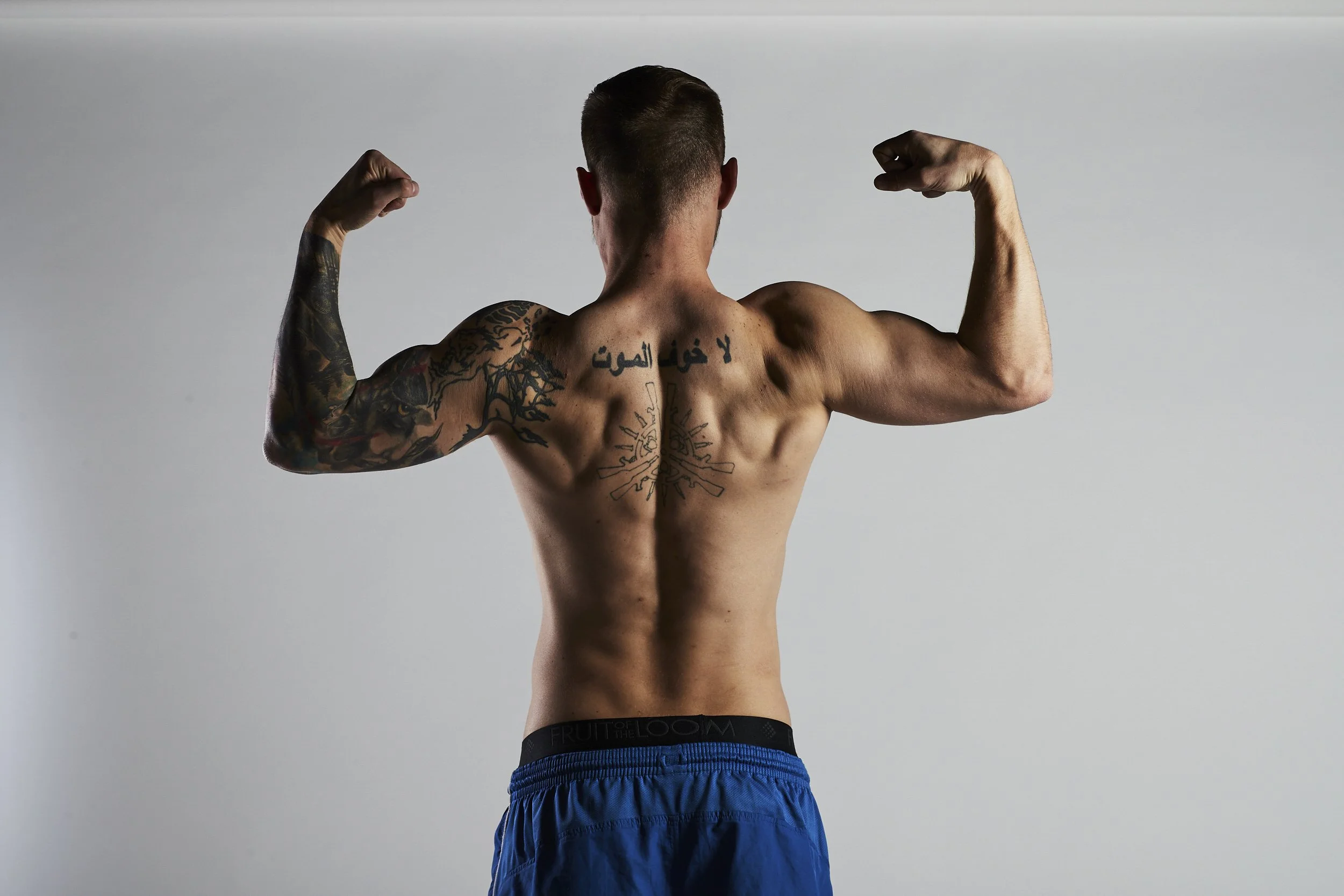 Back view of a shirtless man flexing his biceps, displaying tattoos on his arms and back, wearing blue shorts.