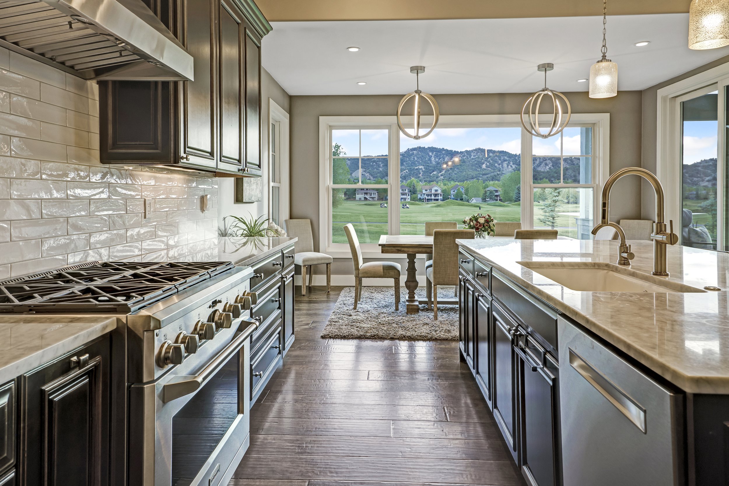 View of a modern kitchen with dark wooden cabinets, beige granite countertops, a stainless steel stove, and a white subway tile backsplash. Large windows provide a scenic view of a green golf course and mountains in the background. There are two hang